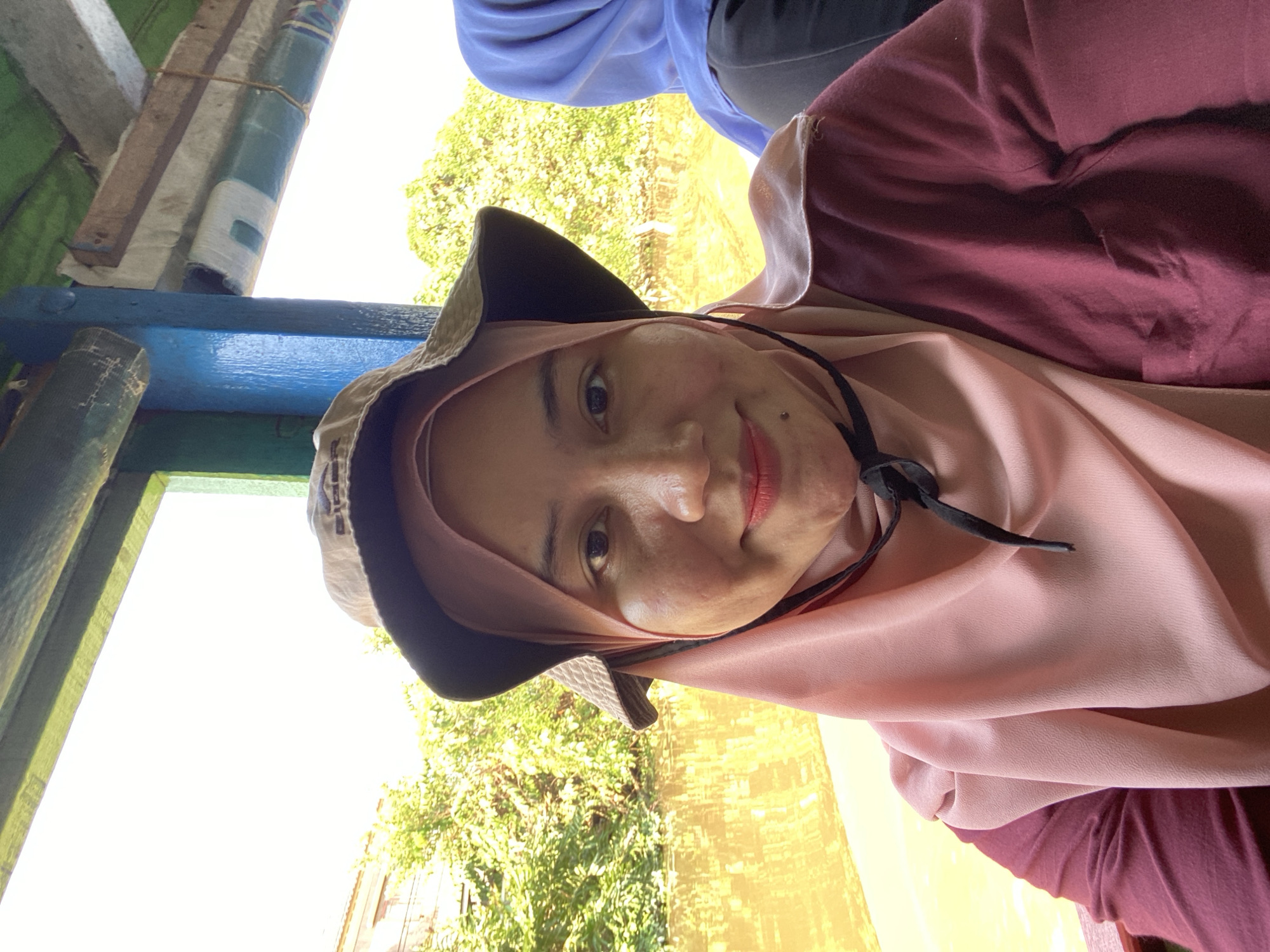 A woman wearing a pink hijab and a maroon long-sleeved shirt is taking a selfie on a boat. She is also wearing a wide-brimmed hat. In the background, there is lush green vegetation and a body of water, likely a river. The boat appears to be covered with a green tarp and has blue and brown structural elements. To the right of the main subject, another person wearing a blue hijab is partially visible. The lighting suggests it is daytime, with bright sunlight. The scene is consistent with locations along the rivers of Banjarmasin, Indonesia, known for its extensive river system and floating markets.