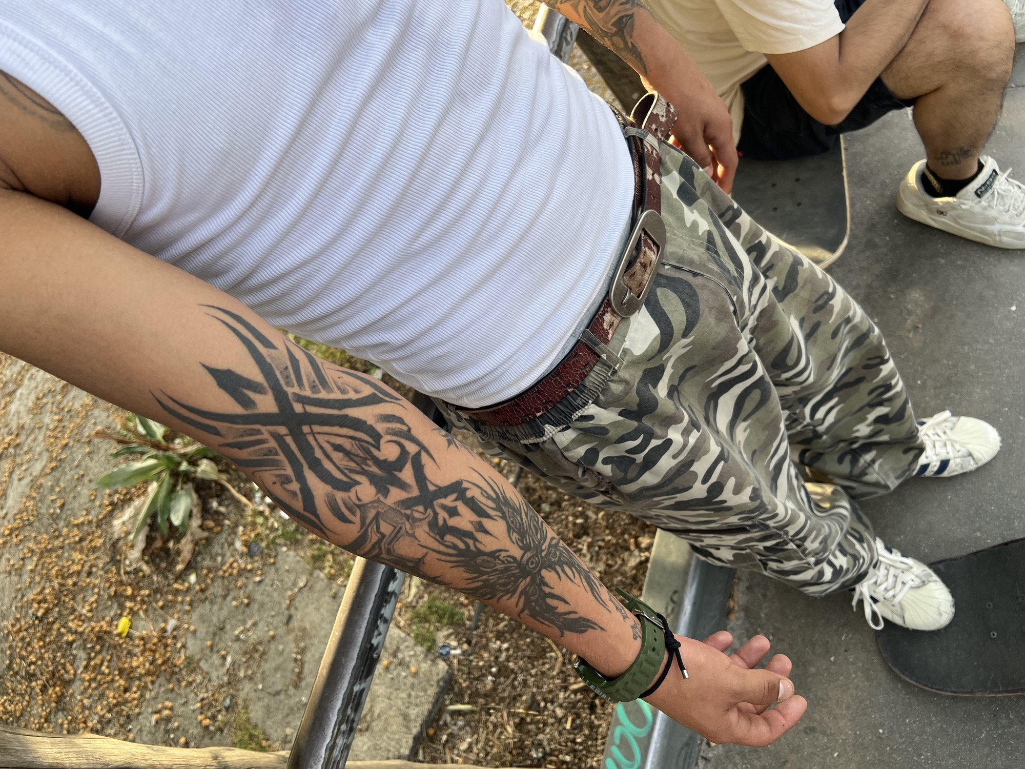 A person with a large black tattoo on their left forearm is shown in an eye-level full shot outdoors. The tattoo extends from the wrist to the bicep and consists of tribal-style patterns and possibly an animal image. The person is wearing a white ribbed tank top, camouflage pants, and a brown belt with a large silver buckle. An olive-green watch is on their left wrist, along with a black bracelet.
To the right of the person is another person sitting on a skateboard. This person is wearing a white shirt and black shorts. Their feet are clad in white Adidas sneakers. Two skateboards are visible on the concrete ground. In the background, there is some foliage and a concrete wall. It is difficult to say definitively if this is Madrid, Spain without further information.