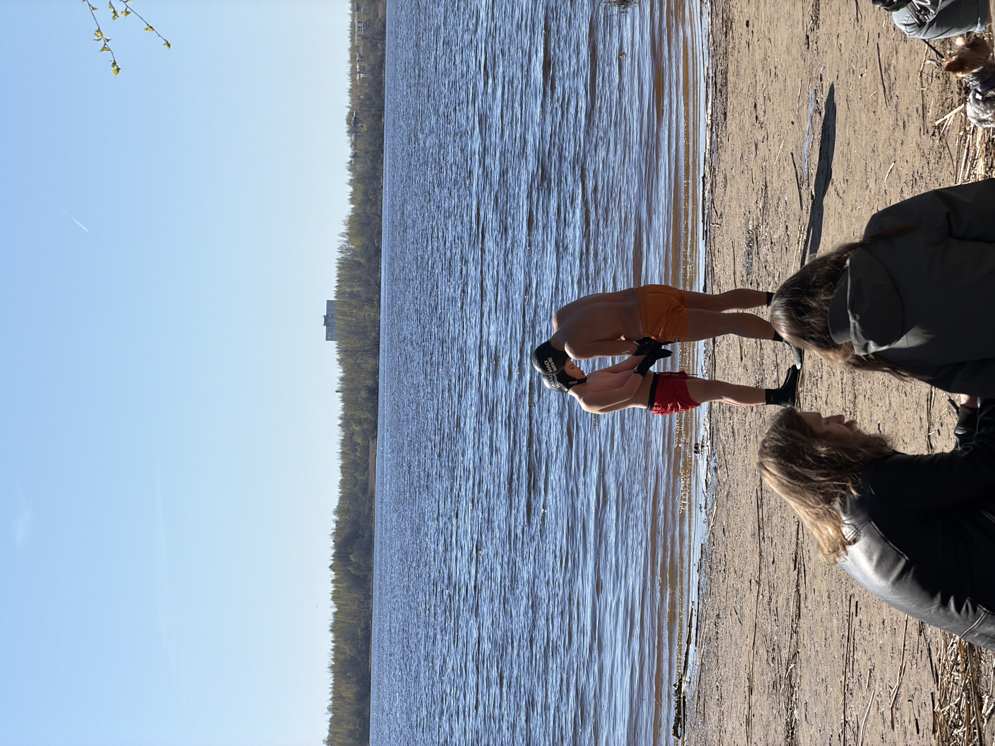 The image shows a sunny beach scene, likely in Saint Petersburg, Russia. Two men in swimwear stand at the water's edge; one wears orange swim trunks and a black swimming cap with white text. The other wears red swim trunks and black socks. Both men appear to be preparing to enter the water. Two people are sitting on the beach watching the men. One is wearing a black leather jacket and the other is wearing a green jacket. A small dog is also present in the scene. The background features a lake or river with trees lining the opposite shore. The sky is clear blue with a single contrail visible. The picture is taken from a high angle in horizontal mode.