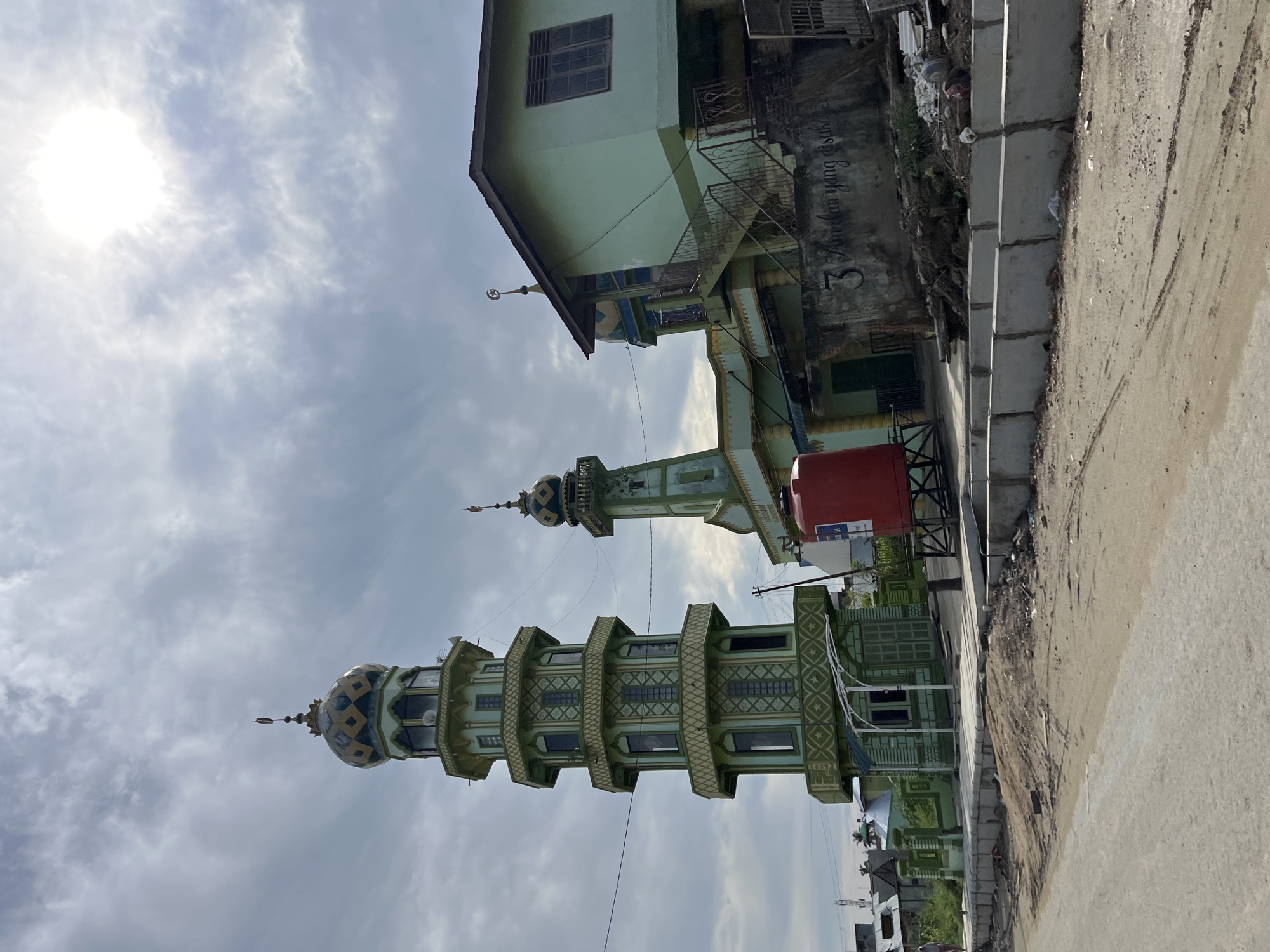 The image depicts a mosque and its minaret in Kota Kuala Simpang, Indonesia. The mosque is a light green structure with yellow accents on its roof and balconies. A tall, slender minaret, also light green with yellow decorative patterns, stands to the left of the mosque. It has multiple levels and is topped with a dome. Another smaller minaret, similar in design, is visible behind the main mosque building. In front of the mosque, a bright red water tank is prominently placed. The ground in front of the mosque is a dusty, unpaved road. A partially constructed concrete barrier runs along the roadside. The sky is overcast with patches of bright sunlight filtering through the clouds. The overall scene suggests a somewhat rural or developing urban environment.