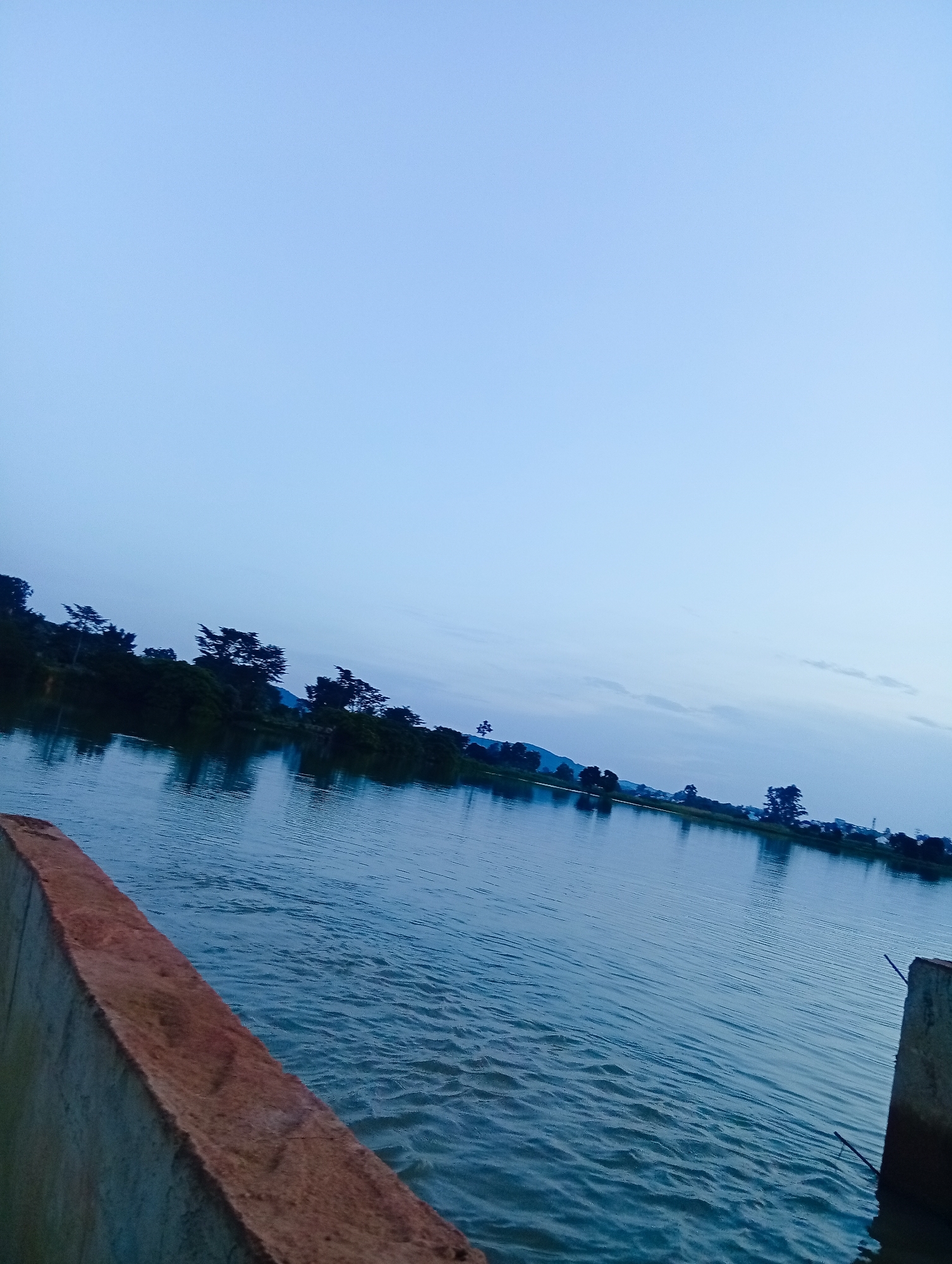 The image depicts a serene, wide body of water, likely a river or lake, under a pale, overcast sky. The water's surface is rippled, indicating a gentle breeze. Along the far bank, a dense line of trees and vegetation forms a dark silhouette against the lighter sky. A few scattered trees are visible further back, suggesting a rural or semi-rural setting. In the foreground, the edge of a concrete structure, possibly a dam or a bridge railing, is visible. The overall atmosphere is tranquil and slightly muted, indicative of early morning or late evening light. This scene is likely located in or around Udu, Nigeria, a region characterized by its natural waterways and lush greenery.
