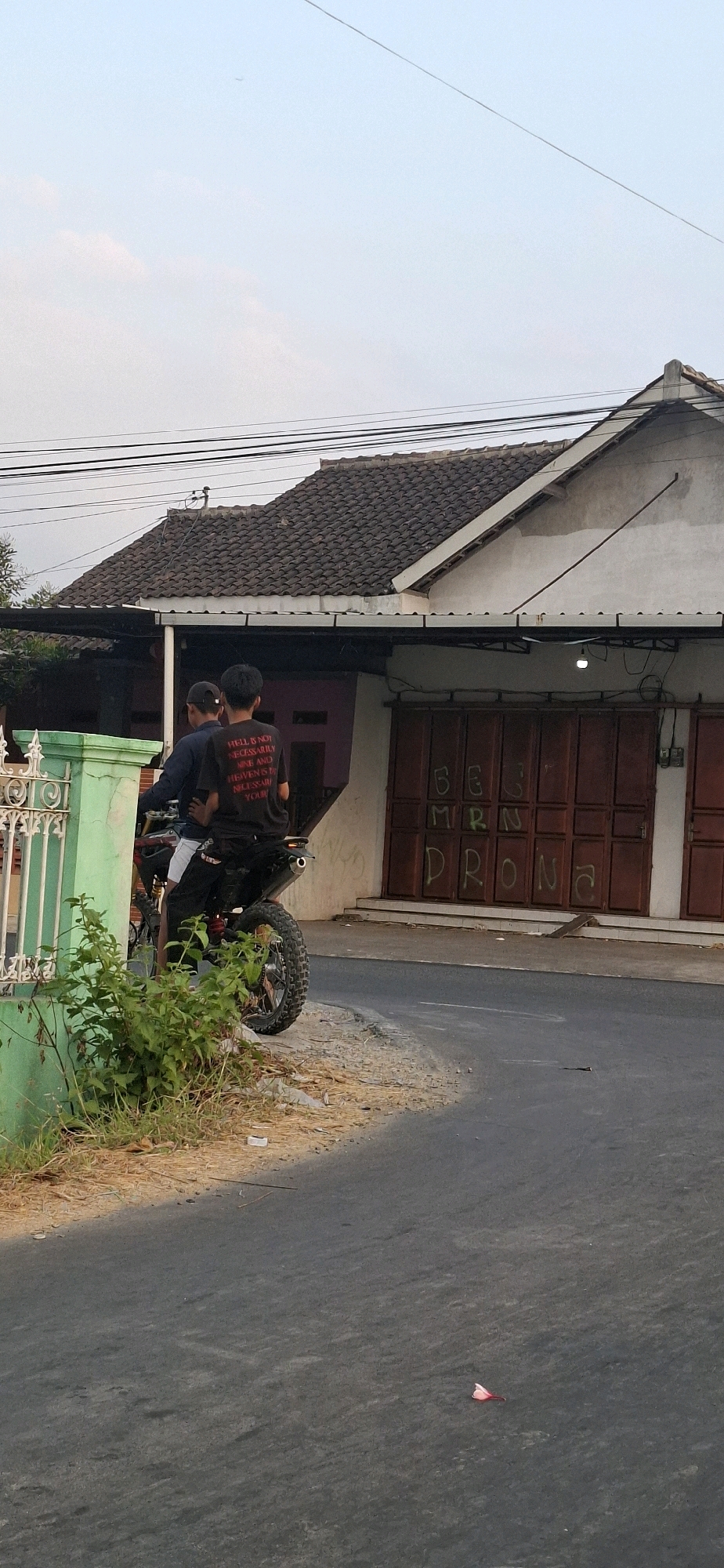 In Desa Dibal, Indonesia, two individuals are positioned on a dirt bike parked on the side of a paved road. The person in the back is wearing a black t-shirt with white text. The bike is facing towards the left side of the image, and the two individuals are looking forward. Behind them, a building with a red corrugated metal door is visible, along with a green fence on the left side of the frame. The sky is light blue with some wispy clouds. The ground is a mixture of asphalt and dry grass, with a few scattered leaves. Overhead, power lines are strung across the frame. The overall scene suggests a casual, possibly a moment of rest or waiting by the side of the road in a rural or semi-urban area.