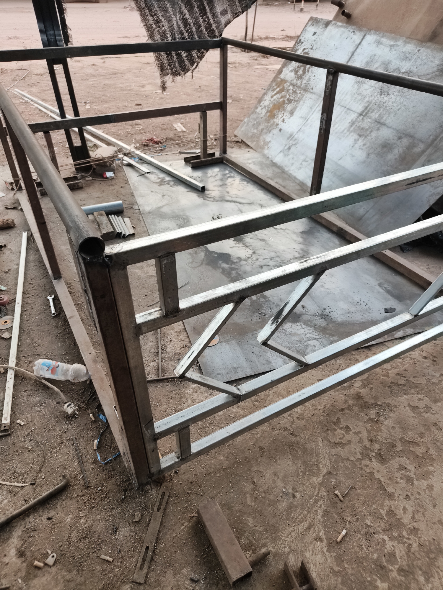 In the city of Suka Harapan, Indonesia, an outdoor metal fabrication site is depicted. The scene shows a partially constructed metal frame, likely for a trailer or platform, lying on the ground. The structure is made of various rectangular metal tubes, some of which are welded together. Metal sheets are visible in the background. The ground is composed of dirt and concrete, with scattered debris, including tools, metal scraps, and a plastic bottle. The overall environment suggests an active workshop area.