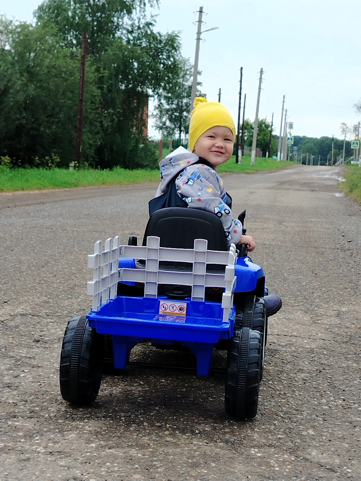 A young child wearing a yellow hat and a grey jacket with car prints is sitting in a blue toy tractor on a grey asphalt road. The child is looking over their left shoulder at the camera and smiling. The tractor has a white fence-like structure at the back. In the background, there are trees, telephone poles, and road signs. The scene appears to be in a rural area or the outskirts of a town.