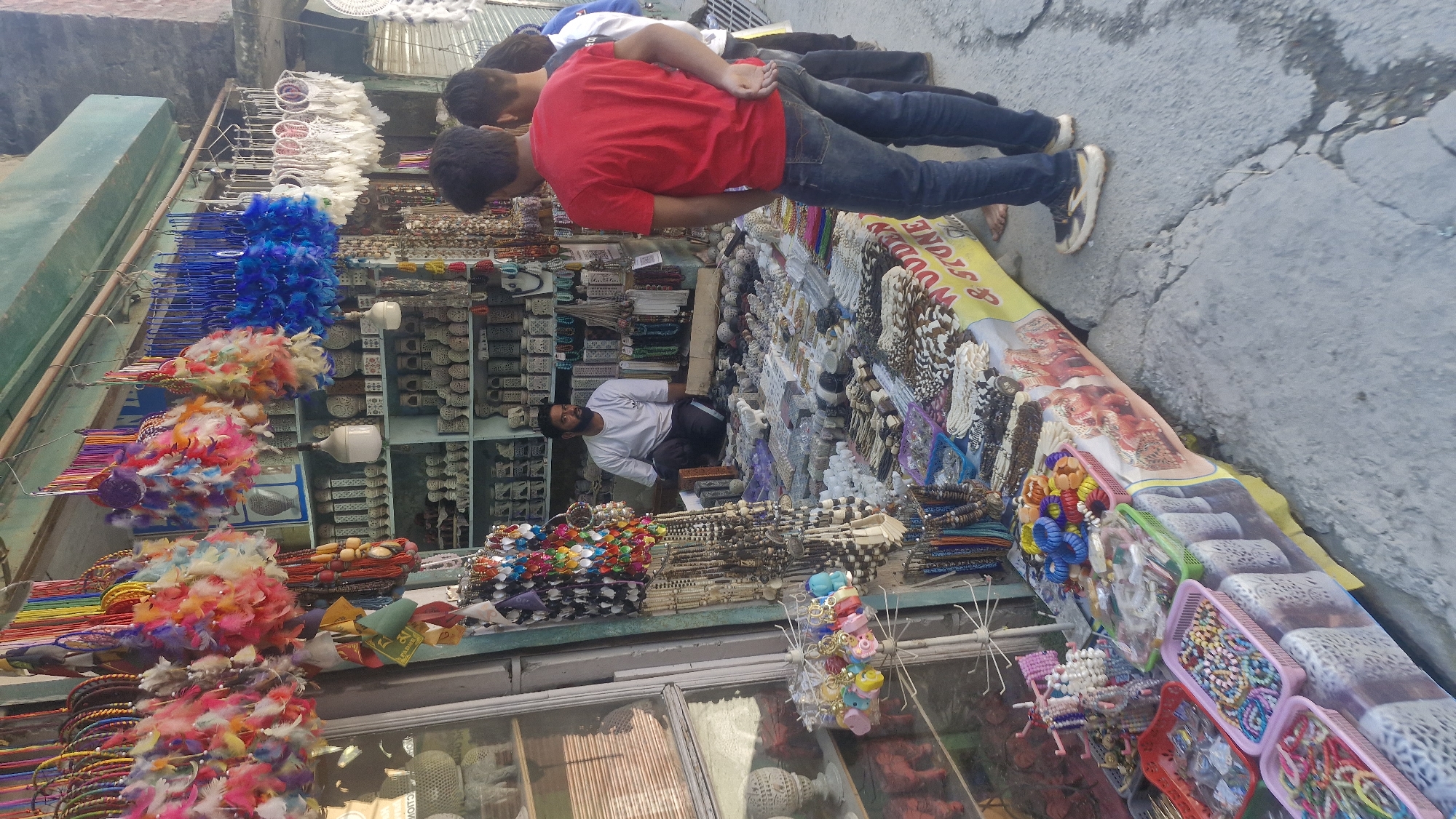 The image depicts a street stall in Vashisht, India, likely a popular tourist area known for its handicrafts and spiritual sites. The stall is laden with a diverse array of items, predominantly jewelry and decorative objects. In the foreground, several plastic baskets overflow with colorful beaded bracelets, necklaces, and other accessories. Behind these, a table displays more intricate beaded items, including necklaces and bracelets made from wood, stone, and other natural materials. The stall itself is covered with a vibrant yellow and red cloth, featuring imagery that appears to be of an Indian theme, possibly deities or mythological figures. Hanging from the stall's canopy are numerous dreamcatchers adorned with feathers in various bright colors, alongside strings of beads and other hanging ornaments.
A vendor, a man with a beard wearing a white t-shirt and dark pants, is seated behind the display, appearing to be attending to customers. In the mid-ground, three young men are observing the merchandise. The man on the right is wearing a red t-shirt and blue jeans, facing away from the camera. Another man stands beside him, partially visible, and a third man is mostly out of frame to the left. The background reveals shelves filled with ceramic or stone decorative items, possibly idols or small sculptures, further suggesting a focus on artisanal crafts. The overall environment is outdoors, with the stall set up on a paved surface, likely a street or a marketplace. The architecture in the background, a stone-like structure, suggests the traditional buildings common in such Indian towns. The lighting indicates it is daytime, with shadows cast on the ground.