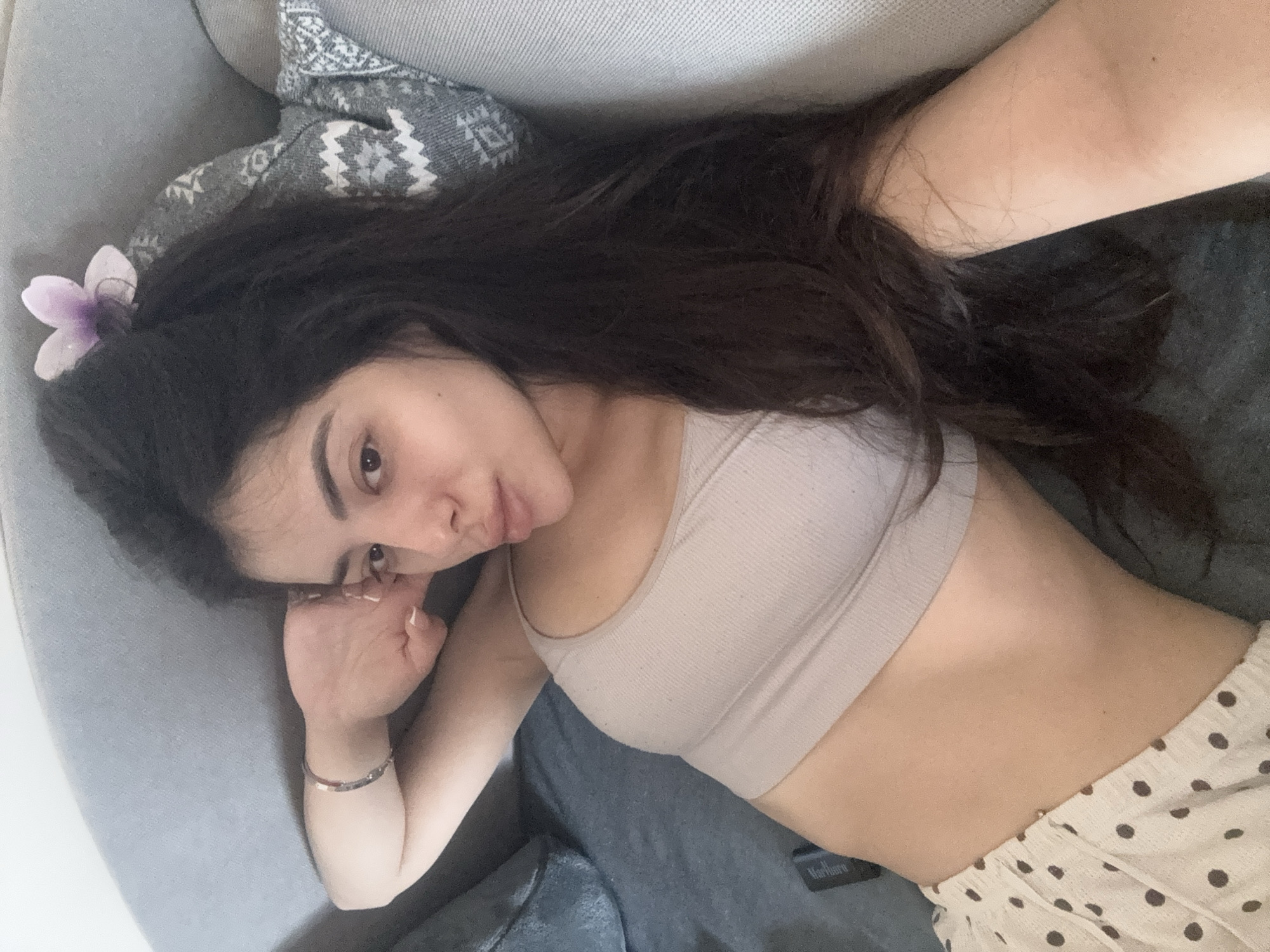 A young woman is captured in a close-up, diagonal shot, lying relaxed on a grey sofa or bed indoors in Plovdiv, Bulgaria. She has long, dark hair, with a small light purple flower clipped near her left temple. Her complexion is light, and she has dark eyebrows, looking directly towards the camera with a calm expression. She is wearing a light beige ribbed bralette or crop top and light-colored shorts with dark polka dots. A thin, reflective bracelet adorns her right wrist, and her nails are neatly manicured white.
She is lying on her right side, propped slightly on her right forearm, with her left arm resting beside her. Behind her head is a grey blanket with a white, patterned design. A dark object, which appears to be a pack of Marlboro cigarettes, is visible near her waist on the grey surface. The word "Marlboro" is visible on this object. The indoor lighting suggests a casual, relaxed time of day.