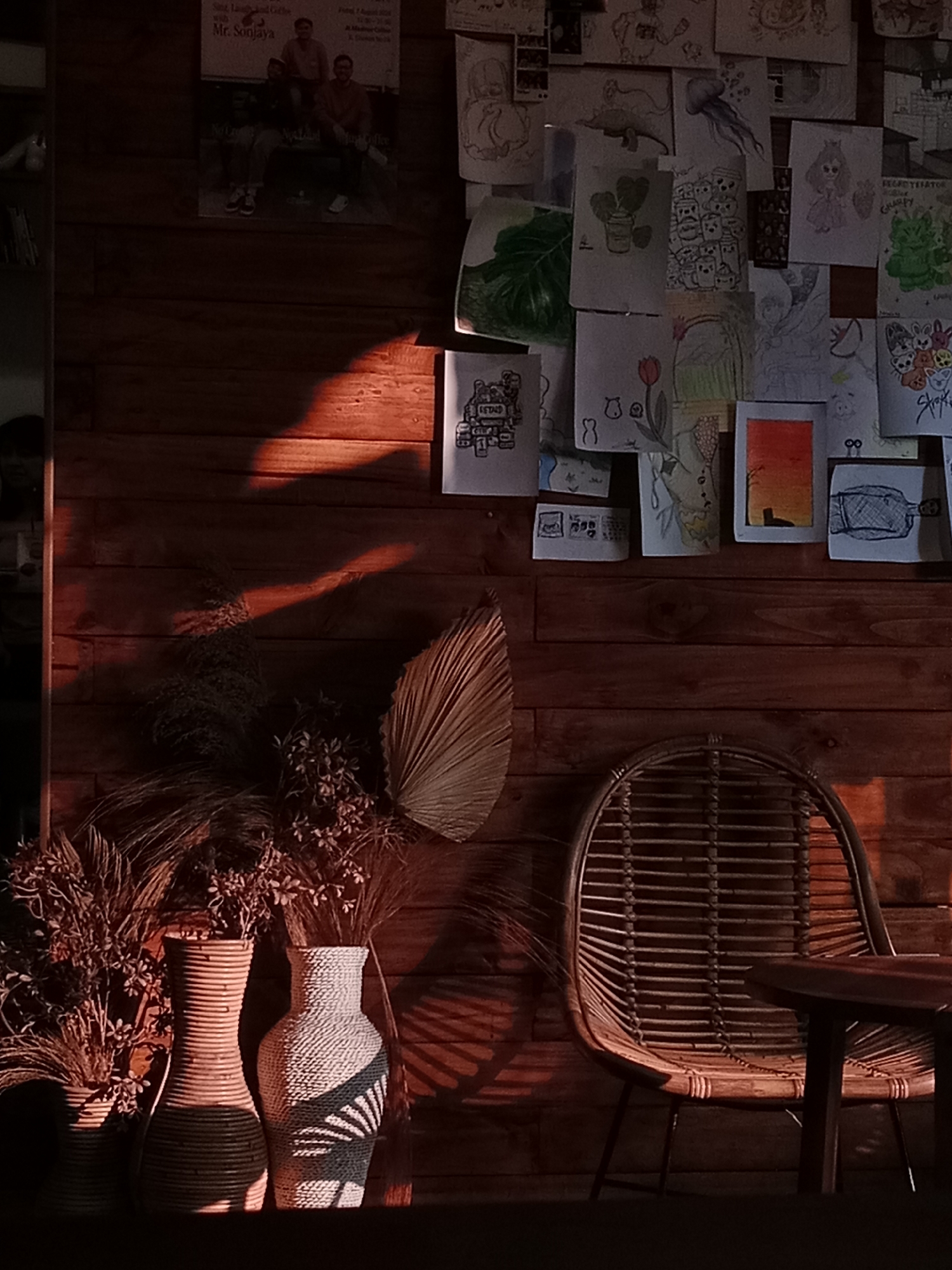The image depicts a dimly lit interior space with warm wooden walls. The wall is adorned with a collage of various drawings and posters, creating a bohemian or artistic atmosphere. In the foreground, there are three decorative vases on a dark surface, two of which are prominently displayed. To the right, a rattan chair with a circular woven seat and back sits beside a small, round wooden table. The lighting suggests late afternoon or early evening, with a strong beam of sunlight casting a dramatic shadow across the wooden wall and floor. The scene appears to be a cozy corner of a cafe or a private residence with a unique aesthetic. There are no people visible in the frame.