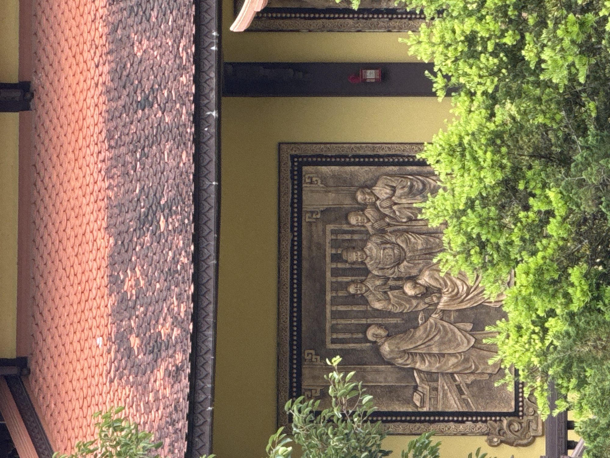 The image shows a section of a building with a red, tiled roof. The wall of the building is painted yellow, and a large, ornate relief sculpture is mounted on it. The sculpture depicts several figures dressed in robes, appearing to be in a formal gathering or ceremony. The figures are rendered in a bas-relief style, with intricate detailing on their clothing and facial features. The scene within the sculpture suggests a hierarchical interaction, with some figures standing and others kneeling. Green foliage from trees is visible in the foreground, partially obscuring the lower portion of the sculpture and the building wall. The architectural style and the relief sculpture suggest a cultural or religious site. This type of architecture and artistry is commonly found in temples and pagodas in Vietnam, including Da Lat.