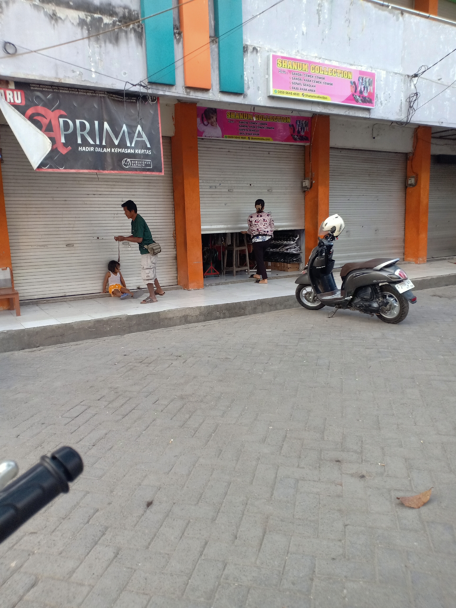The image depicts a street scene in Dander, Indonesia, characterized by a row of shops with closed roll-up doors. In the foreground, a person is visible from the chest down, holding the handlebars of a motorcycle, suggesting they are either about to mount or dismount. A dark-colored scooter with a white helmet on its seat is parked to the right of the frame.
Further into the scene, a man in a green shirt and light-colored shorts stands on the sidewalk, holding a thin rope or string that is attached to a young child. The child, wearing a white tank top and orange shorts, is seated on the ground. To the right of this interaction, a woman in a patterned top and dark pants stands near a shop entrance.
Above the shops, signage indicates "SHANUM COLLECTION" and "PRIMA HADIR DALAM KEMASAN KERTAS." The overall environment appears to be an urban commercial area, with textured paving stones on the sidewalk and road. The buildings have a weathered facade, with visible pipes and wires running along the upper sections. The lighting suggests it is daytime.