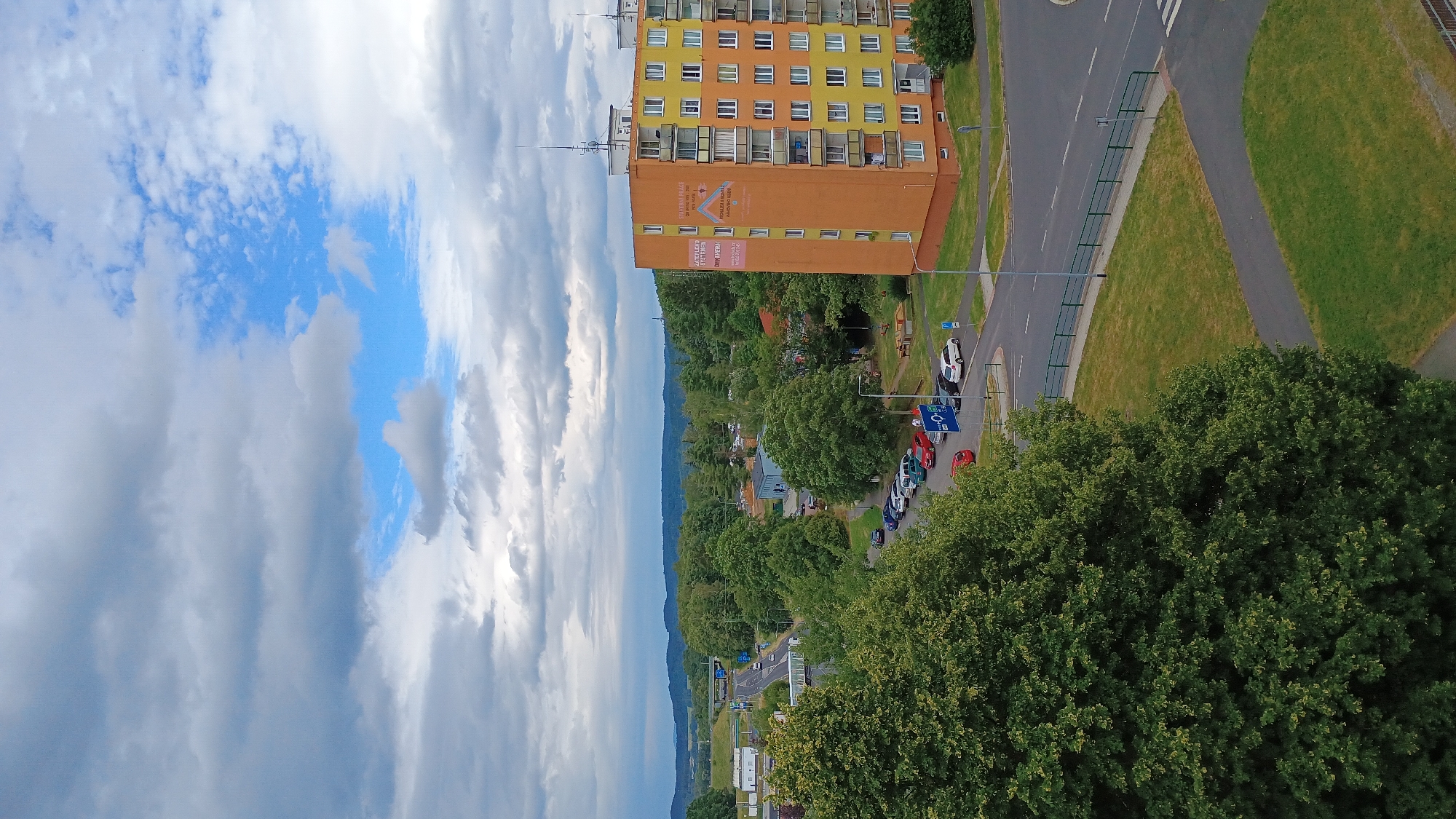 This image captures a view of a residential area in Sokolov, Czechia. In the foreground, a large, leafy green tree dominates the left side of the frame. To the right, a tall, orange and yellow apartment building stands out. The building has multiple floors with balconies and windows. A large banner with text and graphics is affixed to the side of the building.
A street runs in front of the apartment building, with several cars parked along its side. Beyond the parked cars, a road curves into the distance, flanked by more trees and buildings. In the background, rolling hills covered in dense greenery form the horizon, under a sky filled with a mix of dark, heavy clouds and patches of bright blue. The overall scene depicts a typical urban environment interspersed with natural elements.