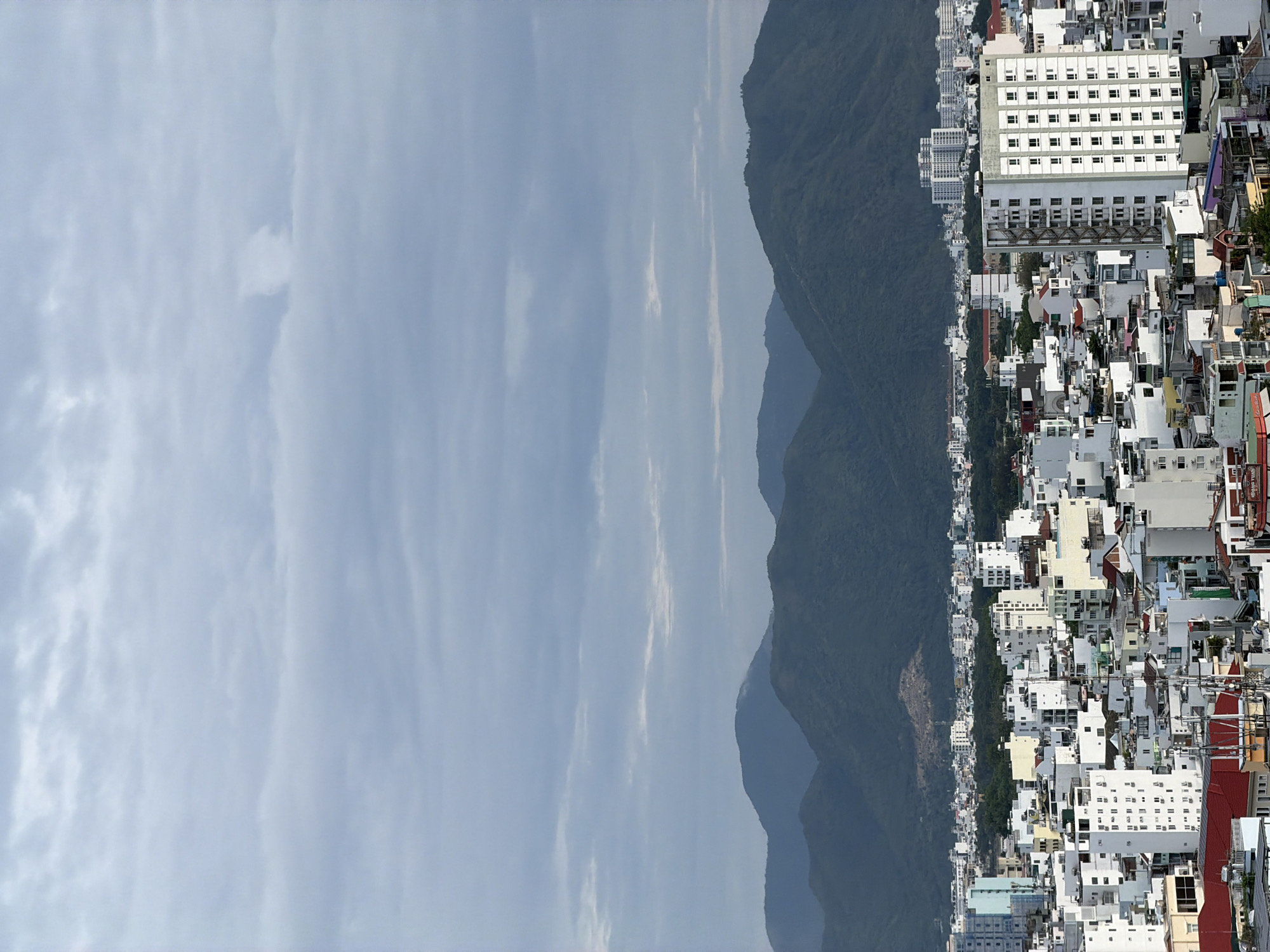 This panoramic landscape image, when viewed in its correct horizontal orientation, depicts a bustling coastal city nestled against a majestic mountain range under a partly cloudy sky. The foreground and midground on the right side of the frame are densely packed with an urban sprawl of buildings. These structures are predominantly white or light-colored, varying in height and design, from multi-story hotels and apartment complexes with numerous windows to smaller residential and commercial units with roofs in shades of red, white, yellow, and grey.
Beyond the urban area, a series of dark green mountains rises prominently, forming a natural boundary across the center of the image. The layered appearance of the mountains suggests depth and distance, their peaks disappearing into a hazy horizon. The left portion of the image is dominated by a pale blue sky dotted with scattered, wispy white clouds, indicating fair weather conditions.
The scene captures a daytime vista, likely late morning or early afternoon, with bright ambient light illuminating the city and natural features. There are no identifiable people or specific activities taking place, presenting a serene yet vibrant overview of the cityscape and its natural surroundings. The overall vista, with its blend of urban density, coastal proximity, and mountainous backdrop, is characteristic of cities like Nha Trang, Vietnam. While some signs are visible on buildings, any text they might contain is largely illegible due to the distance and resolution.