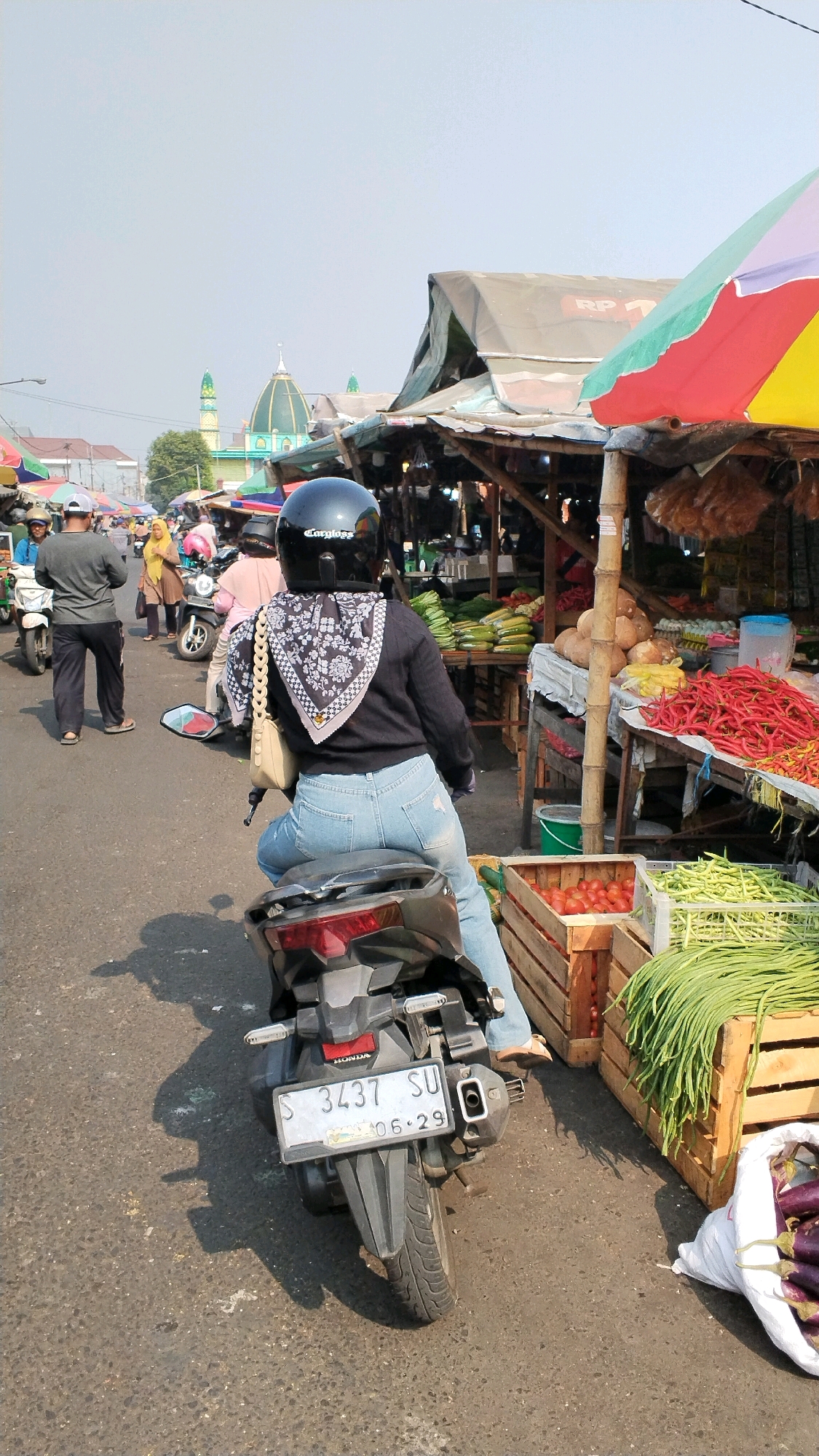 In Mojokerto, Indonesia, a street market scene unfolds under a clear sky. A woman wearing a black helmet and a dark jacket is seated on a black motorcycle, facing away from the camera. She is wearing light blue jeans and has a patterned scarf around her neck and a light-colored purse slung over her shoulder. The motorcycle has a license plate reading "S 3437 SU" and the date "06.29" is visible.
The motorcycle is parked on an asphalt road. To the right of the motorcycle, several stalls are set up selling fresh produce. Wooden crates are filled with long green beans and tomatoes. A pile of red chilies is visible on a table, alongside other vegetables and fruits. In the background, a mosque with a green dome and minarets can be seen, indicating a city environment. Other people and motorcycles are present in the background, suggesting a bustling market. The overall atmosphere is that of a lively outdoor market.