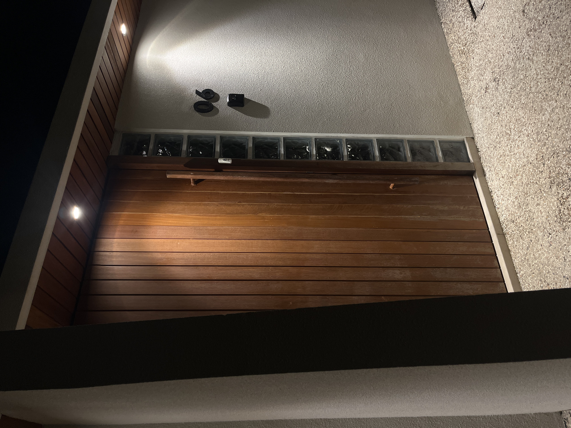 A low-angle, medium shot depicts the facade of a building at night, likely in Kuta Selatan, Indonesia, though specific location identifiers are absent. The primary focal point is a wooden garage door with horizontal planks, positioned beneath a row of translucent, glass-like blocks. Above the glass blocks, the wall is a smooth, light-colored surface. To the left, the building's side is clad with vertical wooden slats. On the wall, two dark, round objects and a black square are visible, casting shadows in the light from an unseen source above. Another light source is present on the left side of the building. In the foreground, a flat surface is present. The overall lighting is a mix of artificial and ambient darkness. The right side of the building features a textured, speckled surface. No people or animals are visible.
