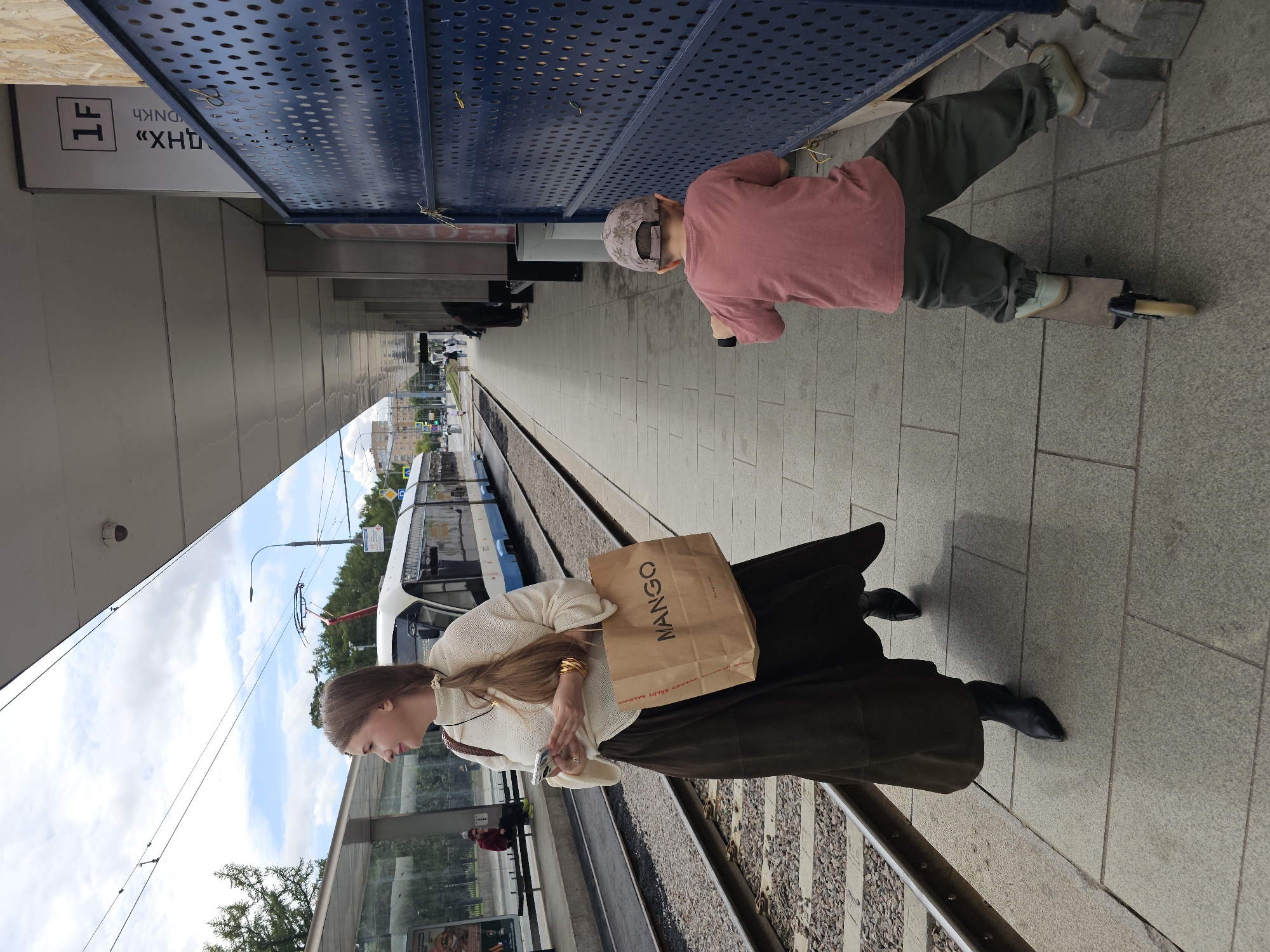 A woman is standing on a tram platform in Moscow, Russia, looking to her left, holding a smartphone and a shopping bag with "MANGO" printed on it. She is wearing a cream-colored sweater and a dark, floor-length skirt with black pointed boots. To her right, a young boy with a cap on backwards is riding a scooter. Behind the woman, a tram is stopped at the station. The platform is paved with stone tiles, and there is a blue perforated metal fence to the right of the boy. Above the fence, a sign displays "1F". The sky is cloudy.