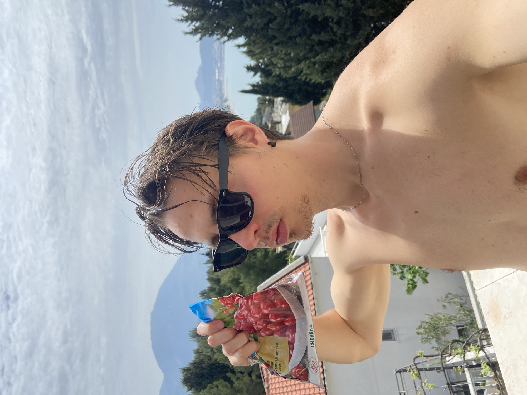 A light-skinned young adult male, with wet, medium-brown hair, wearing black sunglasses and a silver chain necklace, is holding a clear plastic bag of cherries outdoors. The bag reads “CHERRIES” on the front. The man is shirtless. Behind the man, there is a view of buildings, trees, the Adriatic Sea, mountains, and a cloudy sky. The buildings appear to be from the city of Šušanj, Montenegro.