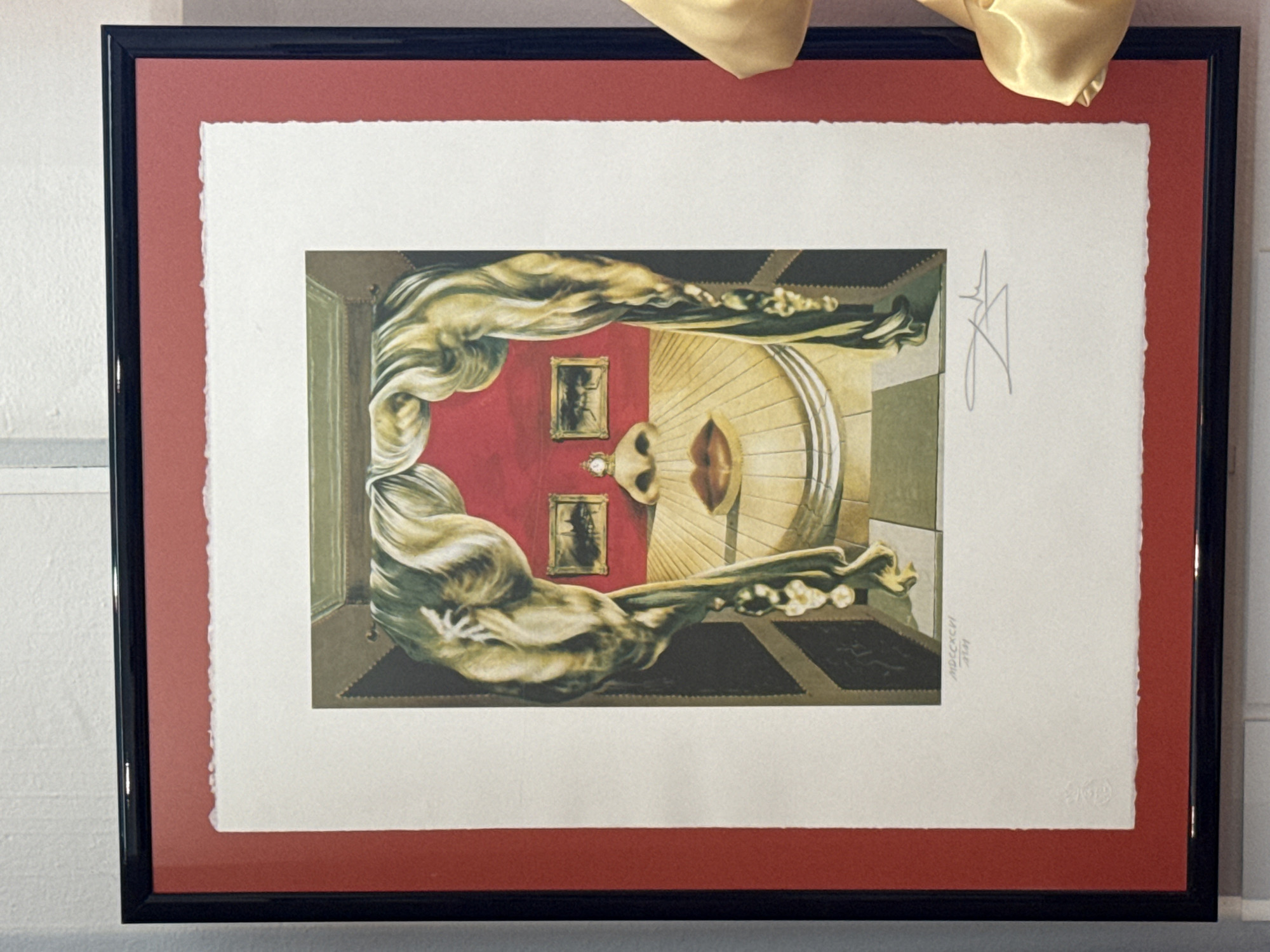 This is a framed print of a surrealist artwork. The artwork depicts a distorted human face within a Renaissance-style room. The face is composed of architectural elements and drapery, with a red background. Two small framed pictures are visible on the red background, and a clock is perched on a nose-like protrusion. The overall style is reminiscent of Salvador Dalí. The print is signed in the lower right corner with a signature and appears to have a date or inscription below the artwork. The print is matted with a rough-edged white border and a red mat, all enclosed in a black frame. The image is captured from a slightly elevated angle, showing the framed artwork hanging on a white wall. There is a hint of a gold fabric draped to the right of the frame, possibly part of a curtain or decoration. The scene is indoors, and the white wall suggests a gallery or exhibition space. The city of Pernik, Bulgaria is not depicted or referenced in the image.