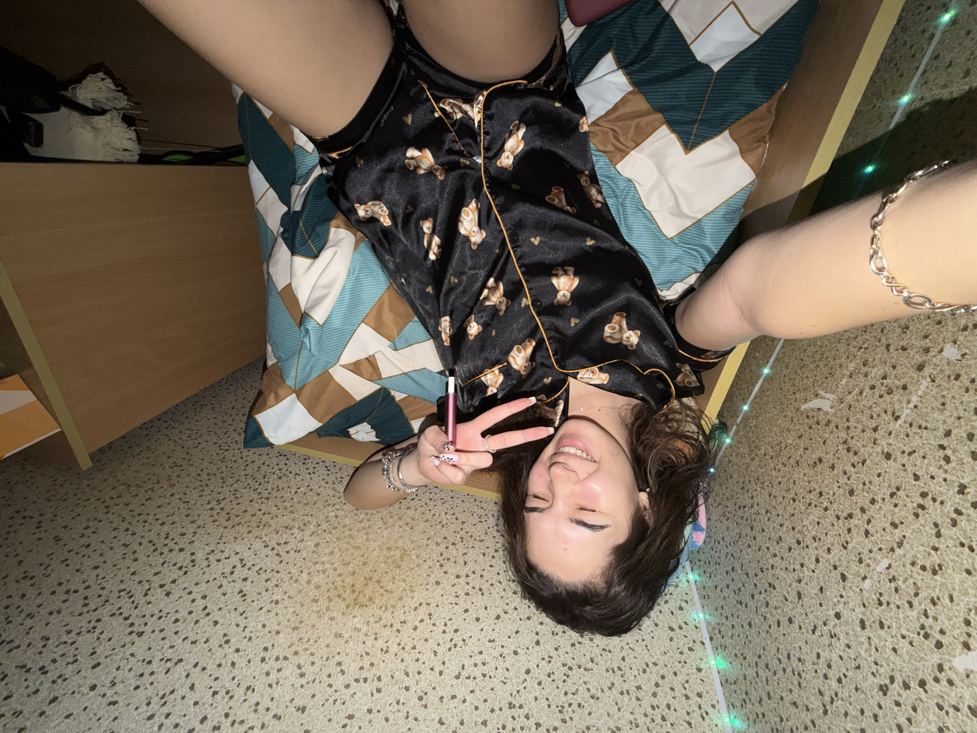 A woman is lying on a bed, smiling and making a peace sign with her left hand, while holding a pink electronic cigarette between her middle and ring fingers. She is wearing black pajamas with a teddy bear print and has bracelets on both wrists. The bed is covered with a quilt with a geometric pattern in shades of green, brown, and white. A bedside table is visible to the left of the bed. The room has a speckled beige floor. The location of the image is not clear from the image provided, and Sofia, Bulgaria cannot be confirmed.
