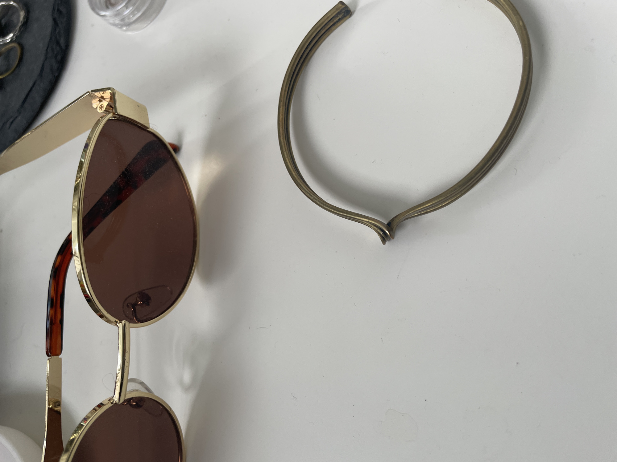 In an indoor setting with a white surface, a pair of gold-rimmed sunglasses with brown lenses and tortoise-shell temples is positioned on the left side of the frame. To the right of the sunglasses, a detached gold-colored metal band, possibly from a bracelet or other accessory, lies on the white surface, curved in a semi-circular shape. In the background, a dark slate-like object with a faint imprint of circular shapes is visible, along with a small clear container. The lighting is bright, suggesting an overhead light source. There are no people or animals present in the scene.