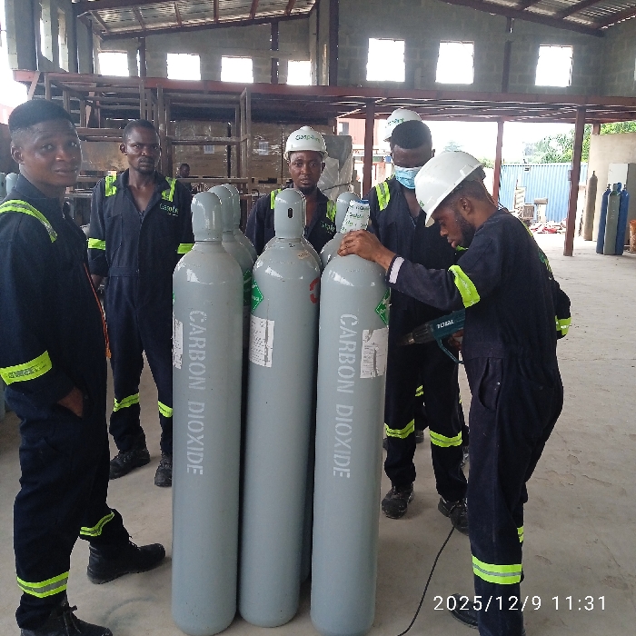 This image, captured on December 9, 2025, at 11:31, depicts five adult males within an industrial facility located in Pakuro, Nigeria. All individuals are dressed in dark blue coveralls featuring yellow-green reflective banding on the sleeves and legs. Three of the subjects wear white hard hats; one hard hat prominently displays the name "Gaspo". The subject on the far right additionally wears a light blue surgical mask.
In the foreground, four tall, upright grey gas cylinders are centrally positioned. Each cylinder is marked vertically with the white text "CARBON DIOXIDE". The individual on the far right is actively holding a black and green "TOTAL" brand heat gun, directing its nozzle towards a label affixed to one of the cylinders, while simultaneously resting his left hand on the cylinder's top cap. The remaining four subjects stand in various positions around the cylinders, some observing the activity, others looking towards the camera.
The environment consists of a concrete floor and a large open space. The background reveals metal structural elements, possibly shelving or equipment frames, and additional gas cylinders. The building features a corrugated metal roof and high-set windows that admit ambient light.