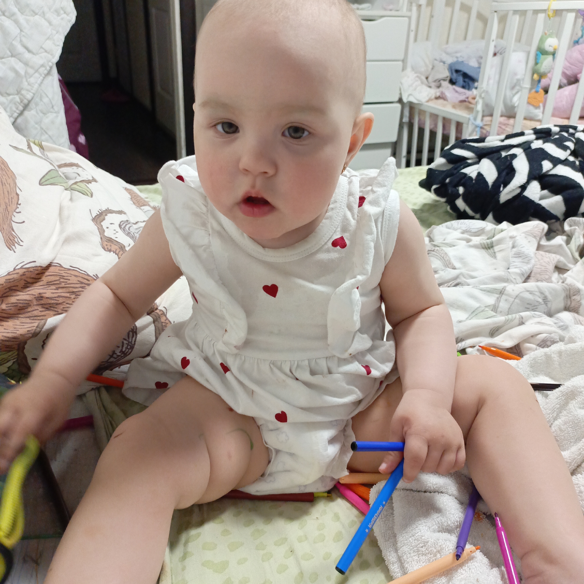 A baby sits amidst a collection of felt-tip pens on a bed with a patterned bedsheet in Unknown, Unknown. The child, wearing a white dress adorned with red hearts, holds a blue pen in their right hand and another object, possibly another pen or toy, in their left. The bedsheets have various designs, including what appears to be a sloth-like animal and a black and white zig-zag pattern. A baby cot is visible in the background, partially obscured by the bed and its contents. The room is cluttered, with additional items lying on the bed and inside the cot.