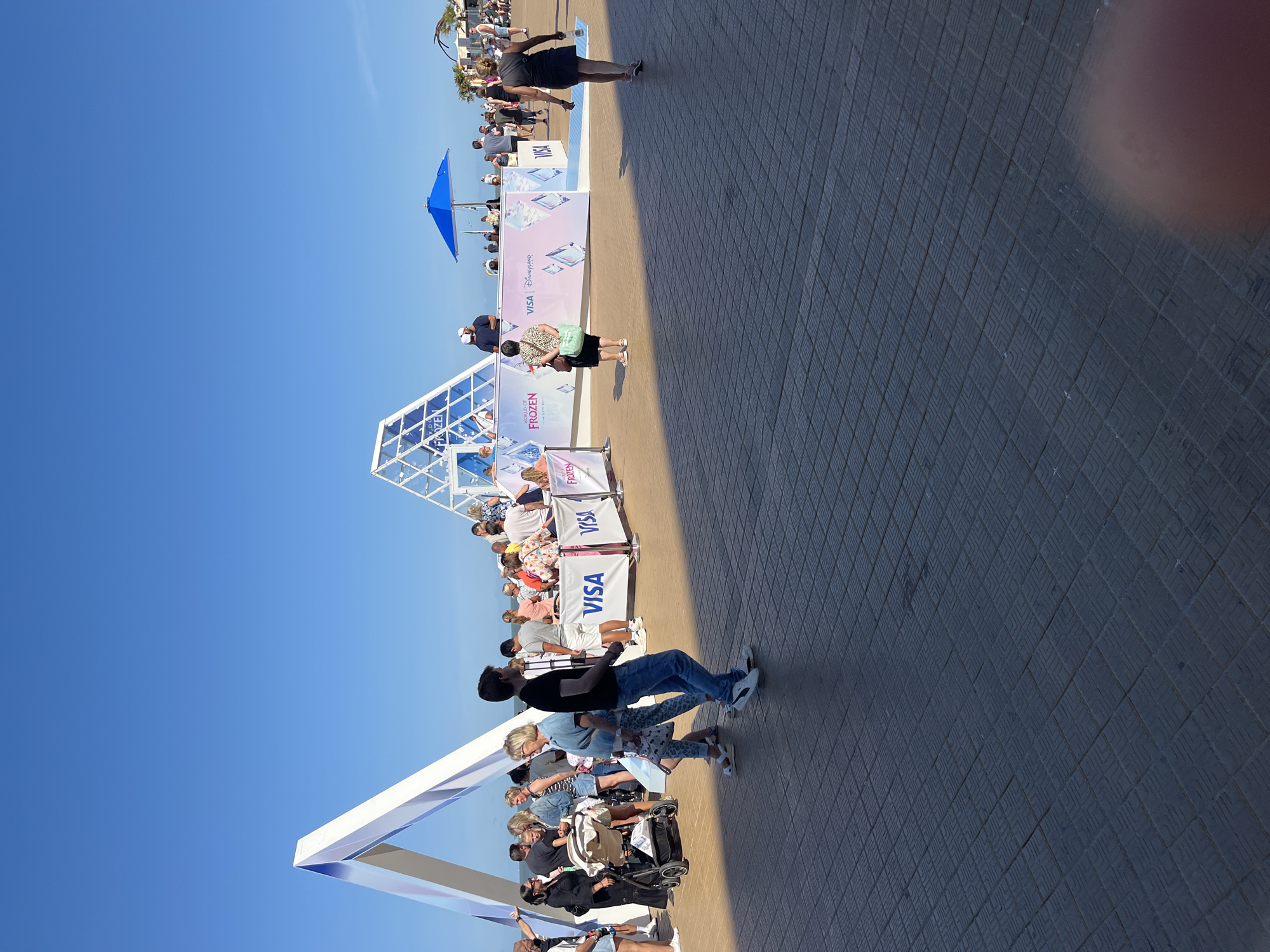 The image depicts a sunny outdoor scene at the Ostend seaside promenade in Belgium. A crowd of people are gathered around a promotional event, likely a temporary setup for "World of Frozen" and "Disneyland Paris," indicated by signage visible on banners and structures. The promotional area features geometric white structures, one in the shape of an A-frame arch and another a faceted pyramid. Banners display the "VISA" logo alongside "FROZEN" and "Disneyland Paris" branding, with stylized diamond graphics.
Several individuals are visible interacting with the event or walking along the promenade. Some are standing and observing, while others are moving through the scene. A stroller is being pushed by a person on the left. In the background, there are palm trees, buildings, and a blue umbrella providing shade. The ground is paved with a patterned dark material. The sky is clear and blue, suggesting good weather conditions. The overall atmosphere appears to be one of a public gathering at a leisure location.