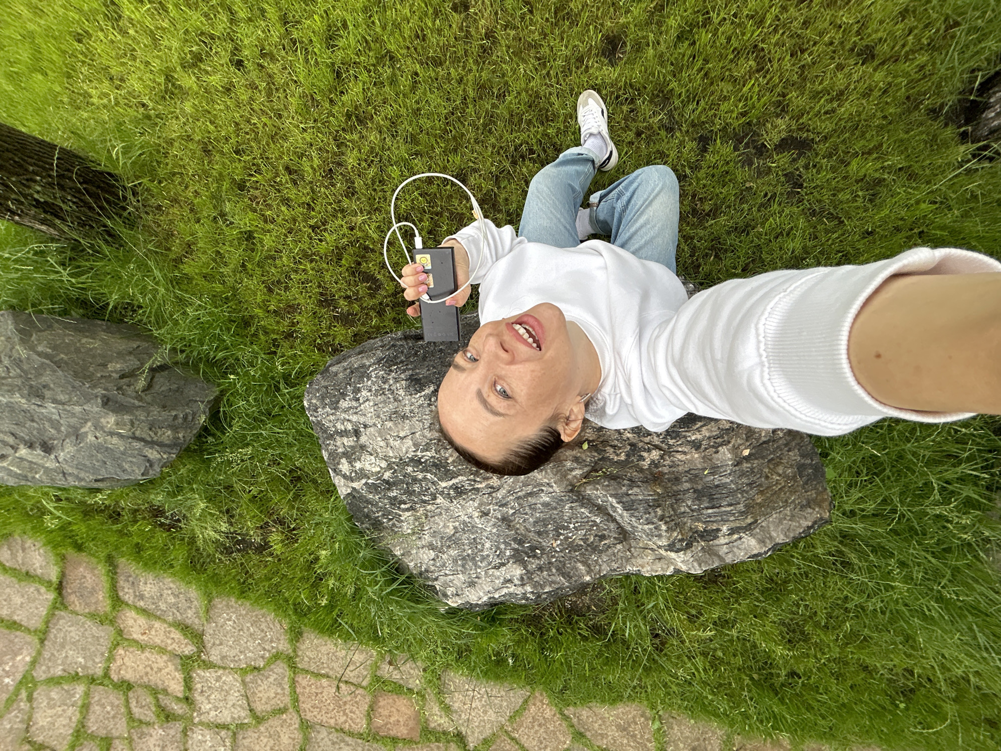 A person is taking a selfie while sitting on a rock in a park. The person is wearing a white sweatshirt, blue jeans, and white sneakers. They are holding a black power bank with a white cable in their left hand. The person is smiling and looking at the camera. The background is a lush green lawn with a paved path and large rocks. The image appears to have been taken during the daytime, with natural light illuminating the scene. The overall mood is cheerful and relaxed. There is no visible text in the image. The location is Yekaterinburg, Russia.