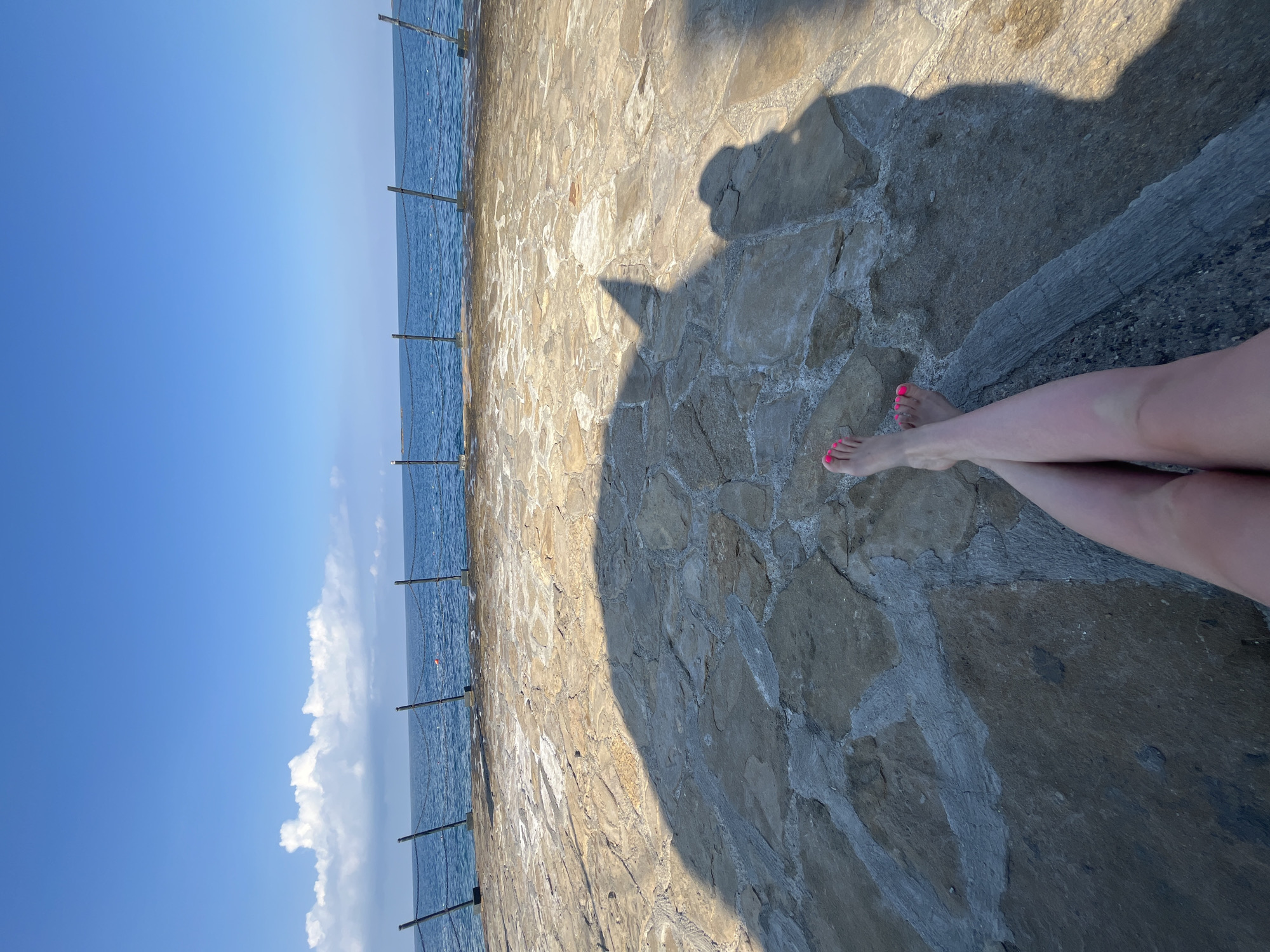 The image shows a person's legs and feet resting on a stone-paved area overlooking the sea. The person's feet are bare, with bright pink toenail polish. A shadow of the person and possibly an object is cast on the paving stones. The sea is blue and calm, with a few small white clouds in the bright blue sky. A chain-link fence with metal posts runs along the edge of the paved area overlooking the sea. The location is identified as Alanya, Turkey.