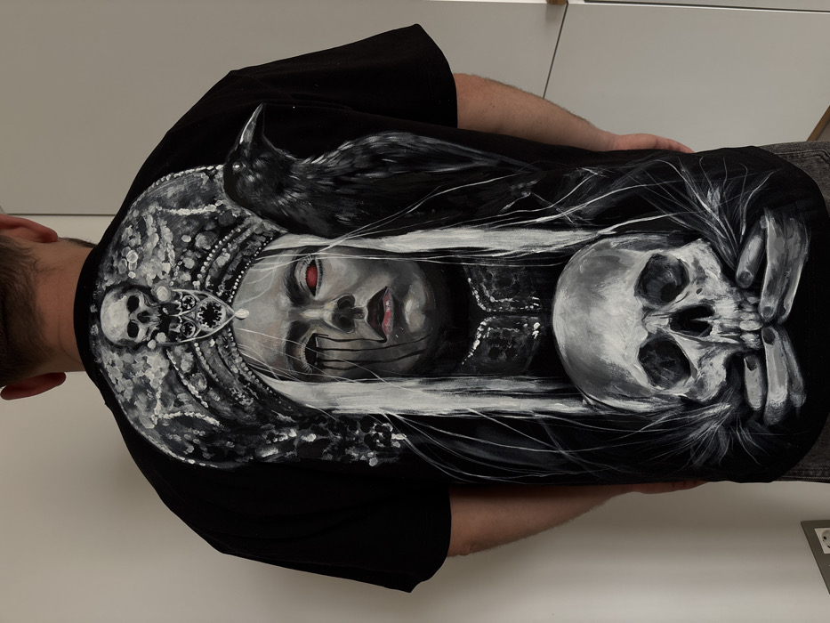 A male individual is observed from a rear perspective, standing indoors against a light-colored wall in Krasnoyarsk, Russia. He wears a black short-sleeved t-shirt featuring a detailed, hand-painted artwork covering the entire back. The artwork depicts a stylized female figure with long white hair, pale skin, red eyes, and dark tears or streaks beneath her eyes, along with red lips. She is adorned with an elaborate, skull-embellished crown. A black raven perches on her right shoulder. Below the female figure, two hands emerge, clasping a human skull. The entire artwork is rendered in a monochrome palette with distinct red accents on the eyes and lips. The individual's short, dark hair is visible at the top of the frame. The immediate environment appears to be a room with plain, light-colored walls and consistent illumination.