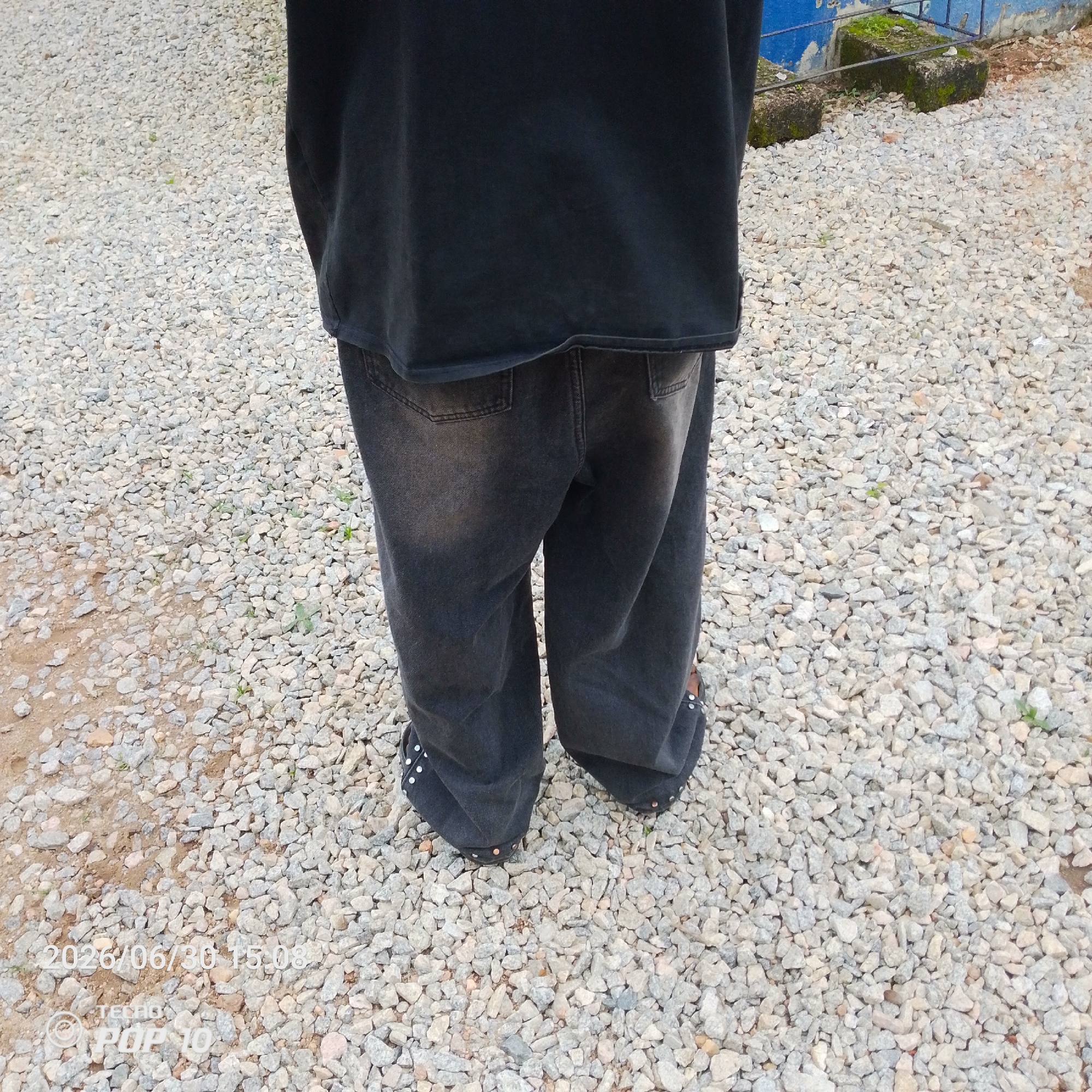 The image shows the lower half of a person wearing a black t-shirt and dark wash, baggy jeans. The person is standing on a surface covered with small, light-colored gravel. The setting appears to be outdoors, possibly a yard or pathway, with some dirt and larger stones mixed in with the gravel. In the background, there are hints of a blue wall with some mossy concrete blocks and a metal railing, suggesting a somewhat dilapidated or rustic environment.
The visible details include the texture of the gravel, the folds and material of the jeans, and the bottom edge of the t-shirt. The person's shoes are visible, appearing to be dark flats with decorative studs along the edges. The lighting suggests it is daytime, but the specific time of day is further clarified by the text overlay.
The overlaid text in the lower left corner reads "2026/06/30 15:08" and "TECNO POP 10". This indicates the date and time the photo was taken, as well as the model of the phone used. There is no clear activity or event taking place, as the person is simply standing still. The image is focused on the lower body and the ground, with no visible facial expressions or emotional cues. The location context provided is Jalingo, Nigeria, which aligns with the outdoor, possibly semi-arid or tropical, environment suggested by the gravel and vegetation.
