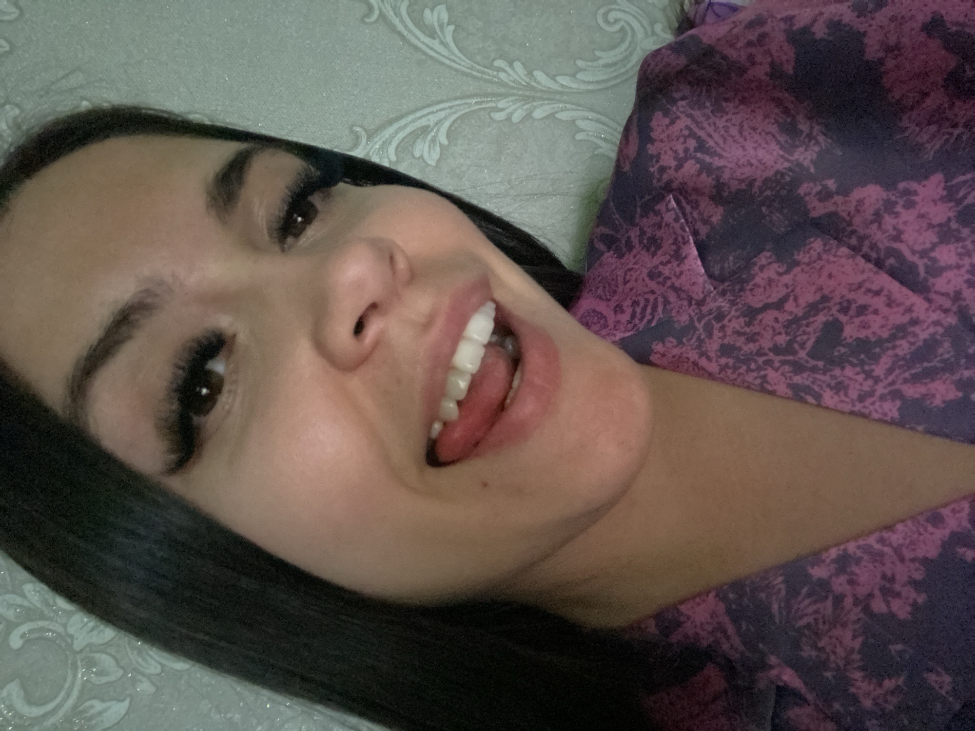 A woman with dark hair, fair skin, and long eyelashes is smiling with her mouth open, exposing her tongue and teeth. She is wearing a purple and pink floral print top. The background appears to be a light-colored wall with a decorative pattern. There is no indication of this picture being taken in the city of Chaikovsky, Russia.