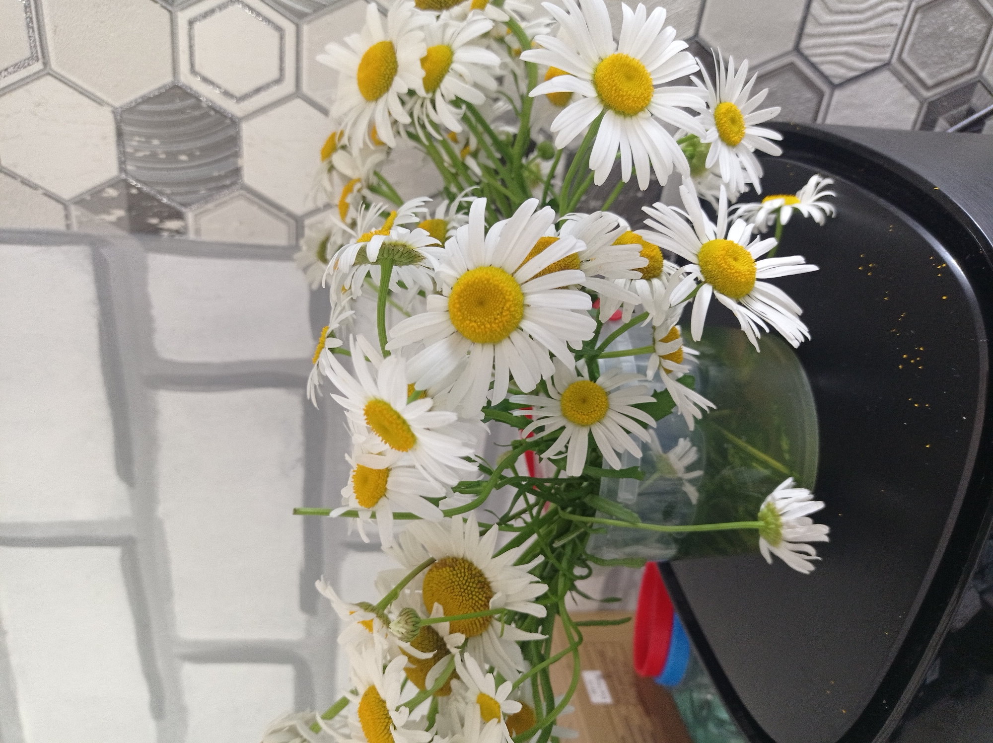 A bouquet of white daisies with yellow centers is displayed in a clear glass vase. The vase sits on a black surface, which appears to be a table. The background consists of a tiled wall, with one section featuring a brick pattern and the other a hexagonal pattern with grey and white tones. In the lower left portion of the image, a red and blue object is partially visible. The overall lighting suggests an indoor setting. There are no people or animals present in the image. The setting does not provide specific clues to pinpoint a location within Uglekamensk, Russia.