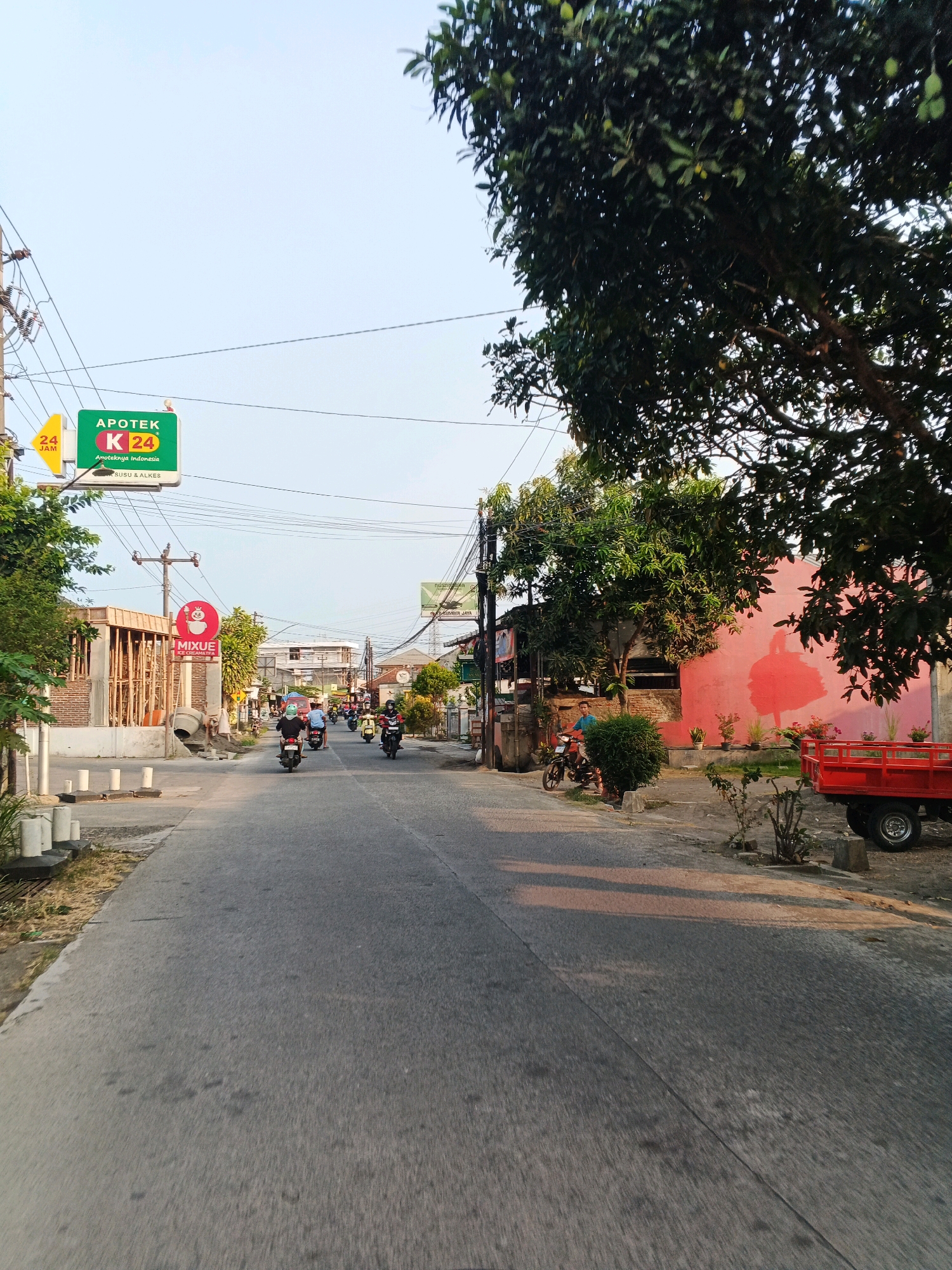 The image depicts a street scene in Planjan, Indonesia, likely during late afternoon, given the long shadows cast across the asphalt. The road is paved and appears to be a local thoroughfare with moderate traffic, primarily consisting of motorcycles. On the left side of the road, there is an ongoing construction project with visible brick walls and wooden framing. A yellow sign with "24 JAM" and the logo "APOTEK K 24" indicates a 24-hour pharmacy. Further down the street, a red sign with a white character and the word "MIXUE" suggests a commercial establishment.
The right side of the street features a vibrant pink building with a large silhouette of a bird or animal painted on its exterior. Adjacent to this building is a bright red flatbed trailer attached to a small vehicle. Lush green trees with visible leaves and some fruit are prevalent throughout the scene, with a large tree dominating the foreground on the right, its branches extending into the upper right corner of the frame. The sky is clear and light blue, suggesting fair weather. Several individuals are seen riding motorcycles, navigating the street. The overall impression is of a typical day in a developing urban or semi-urban area in Indonesia.