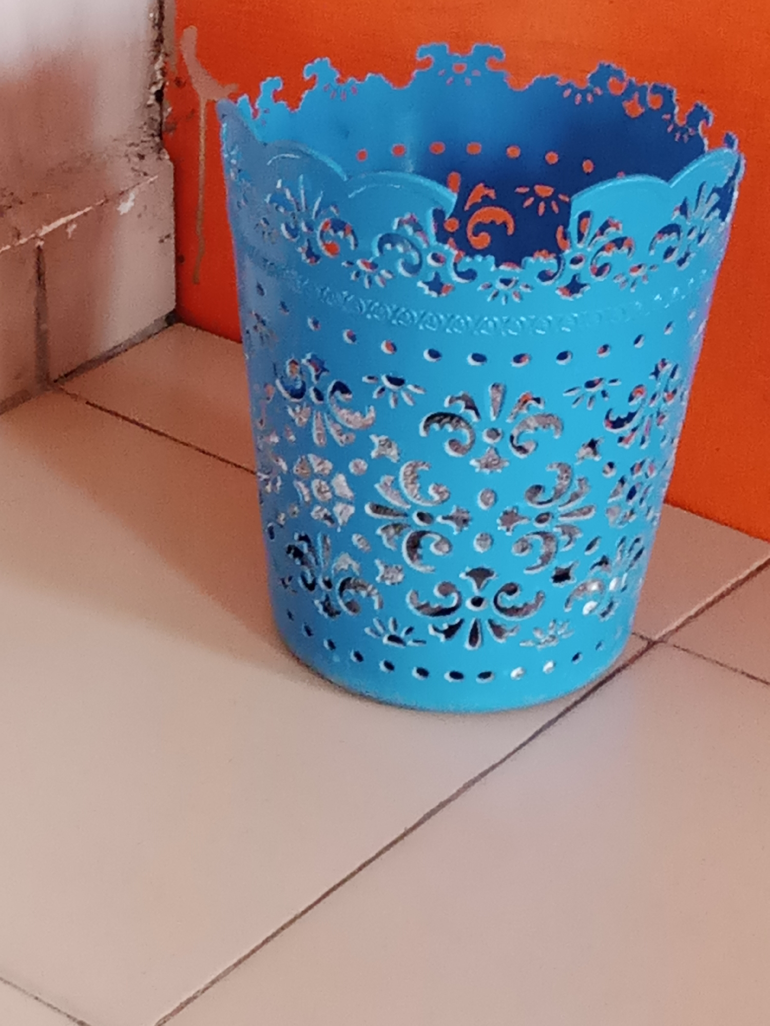 In Jalingo, Nigeria, a blue decorative waste bin with intricate cut-out patterns stands on a tiled floor. The bin has a scalloped edge and is adorned with floral and circular designs, revealing an orange interior. To the left of the bin, a wall with a rough, textured surface is visible, juxtaposed with a section of smooth, lighter-colored tiles. The background features a vibrant orange wall. The scene is static, with no individuals or actions depicted. The lighting suggests an indoor environment, possibly a room or a corner of a building.
