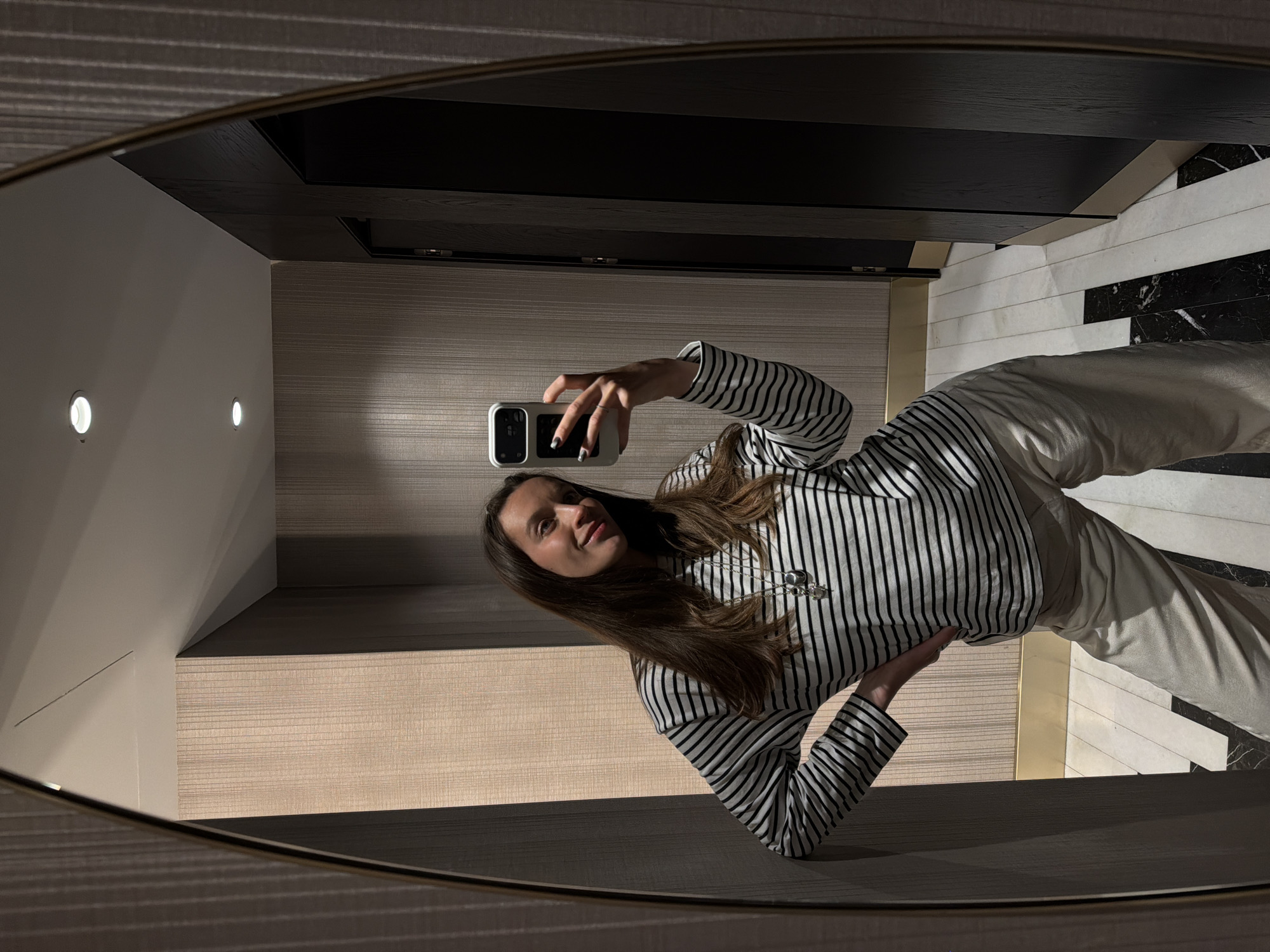 A woman is taking a mirror selfie in what appears to be a hotel room or apartment bathroom. She is holding a smartphone and wearing a striped long-sleeved shirt and light-colored pants. The bathroom features a black countertop or shelf above, with horizontally patterned wallpaper or panels below. The floor is tiled in a black and white geometric pattern. The mirror has a distinctive oval shape and a metallic frame.
The location is difficult to determine definitively from the image alone, but given the question's prompt, it is in Budva, Montenegro. The specific establishment cannot be identified without further visual or contextual information.