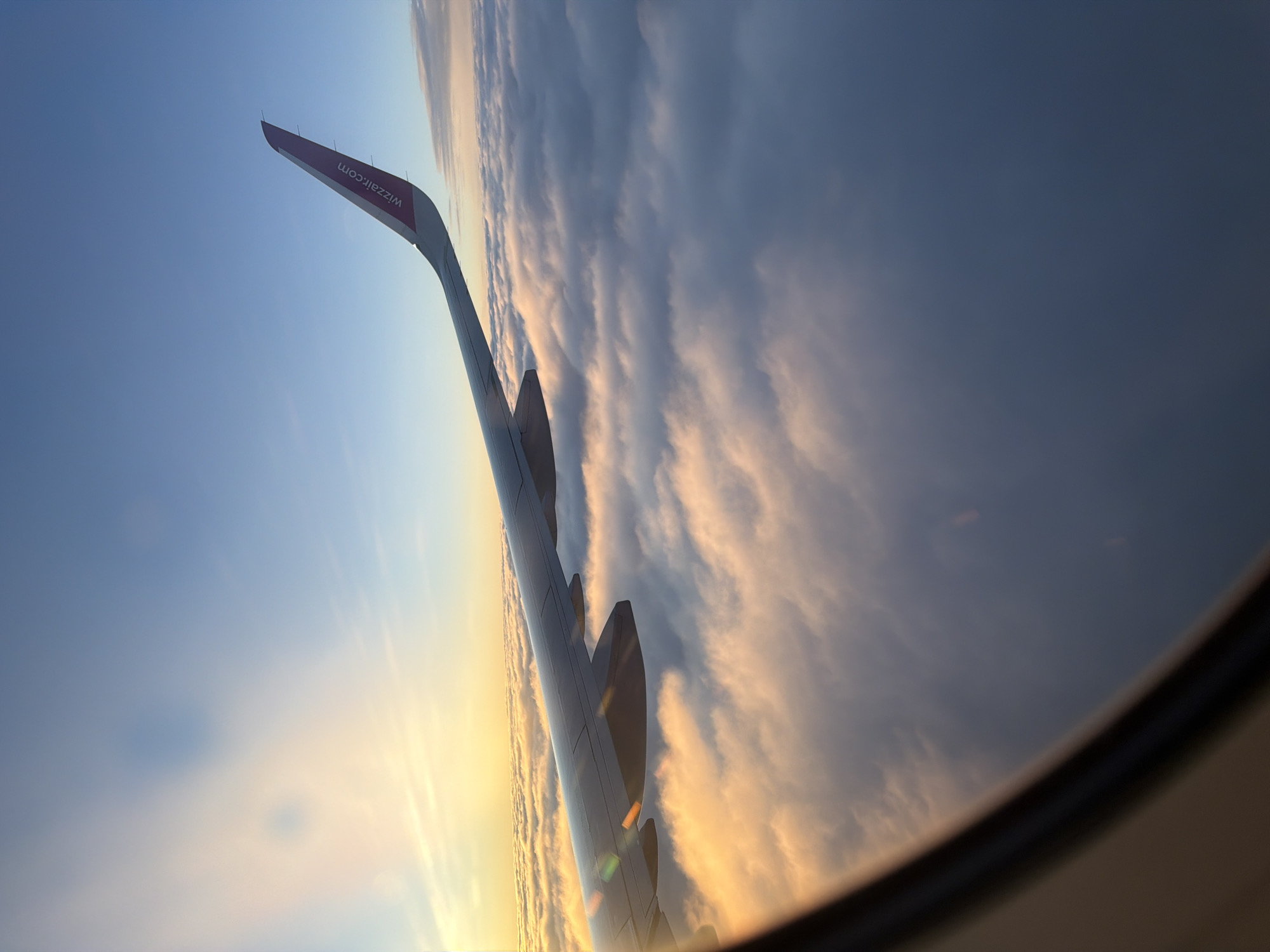 The image is taken from an airplane window, showing an airplane wing extending across the frame. The wing is a dark gray color and has the "wizzair.com" logo in pink on its tip. Below the wing, a thick layer of clouds stretches to the horizon. The sky above is a gradient of blue, transitioning to a warm yellow and orange near the horizon, indicating either sunrise or sunset. The sun is obscured by clouds, but its light illuminates the clouds from behind, creating a soft glow. There are no people visible in the image. The environment is that of being in an airplane flying above the clouds. The location in the city of Czerwony Kościół, Poland is not discernible from the image itself.
