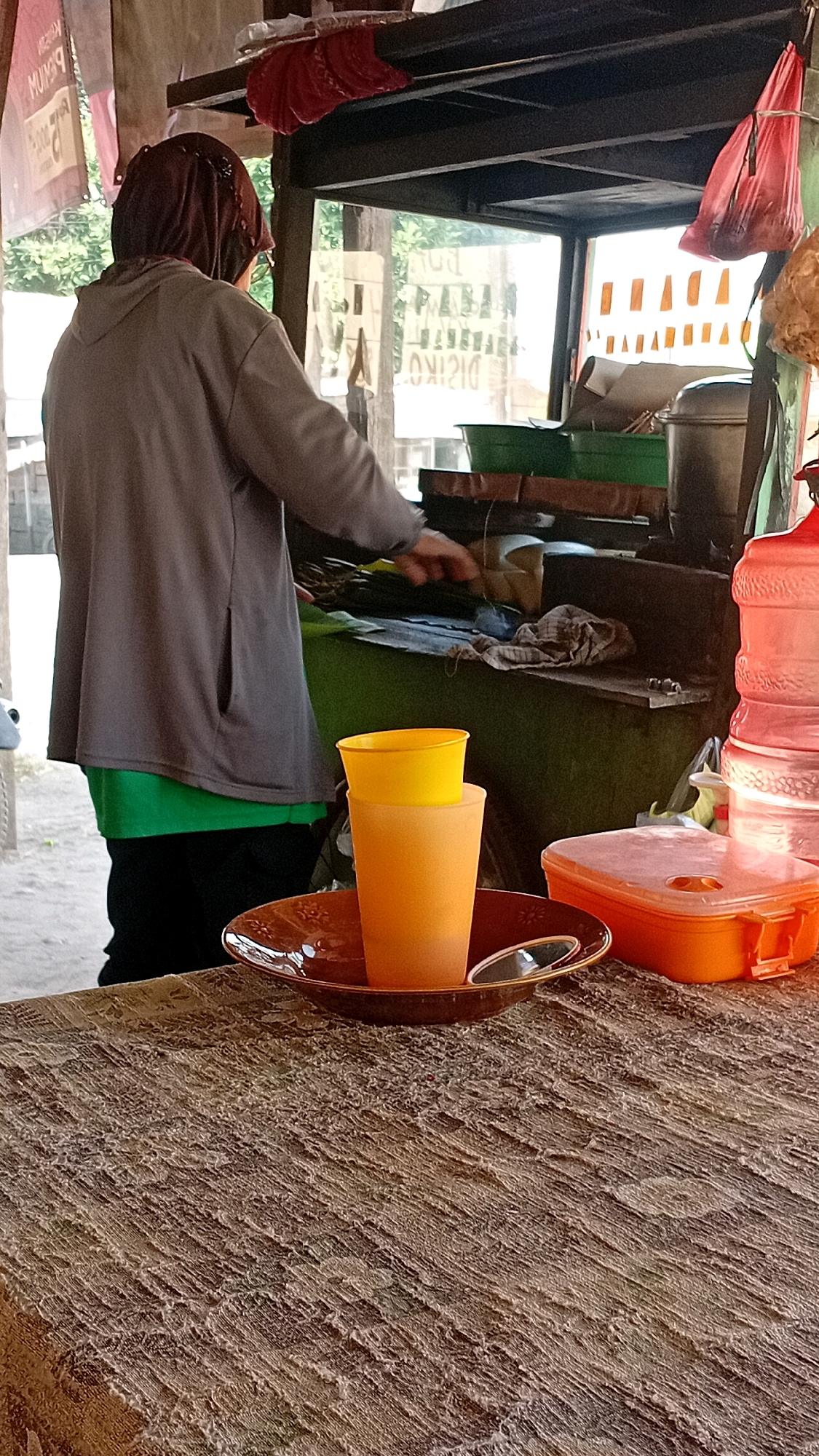 A person wearing a brown hijab and a grey hooded jacket is standing behind a food stall. The stall has a metal frame and shelves, with various items displayed including plastic containers, a large water jug, and what appear to be food items wrapped in leaves. In the foreground, a brown decorative plate holds two plastic cups, one orange and one yellow. The table in front of the stall is covered with a patterned brown tablecloth. The background shows foliage and buildings, suggesting an outdoor urban setting in Jambi, Indonesia.