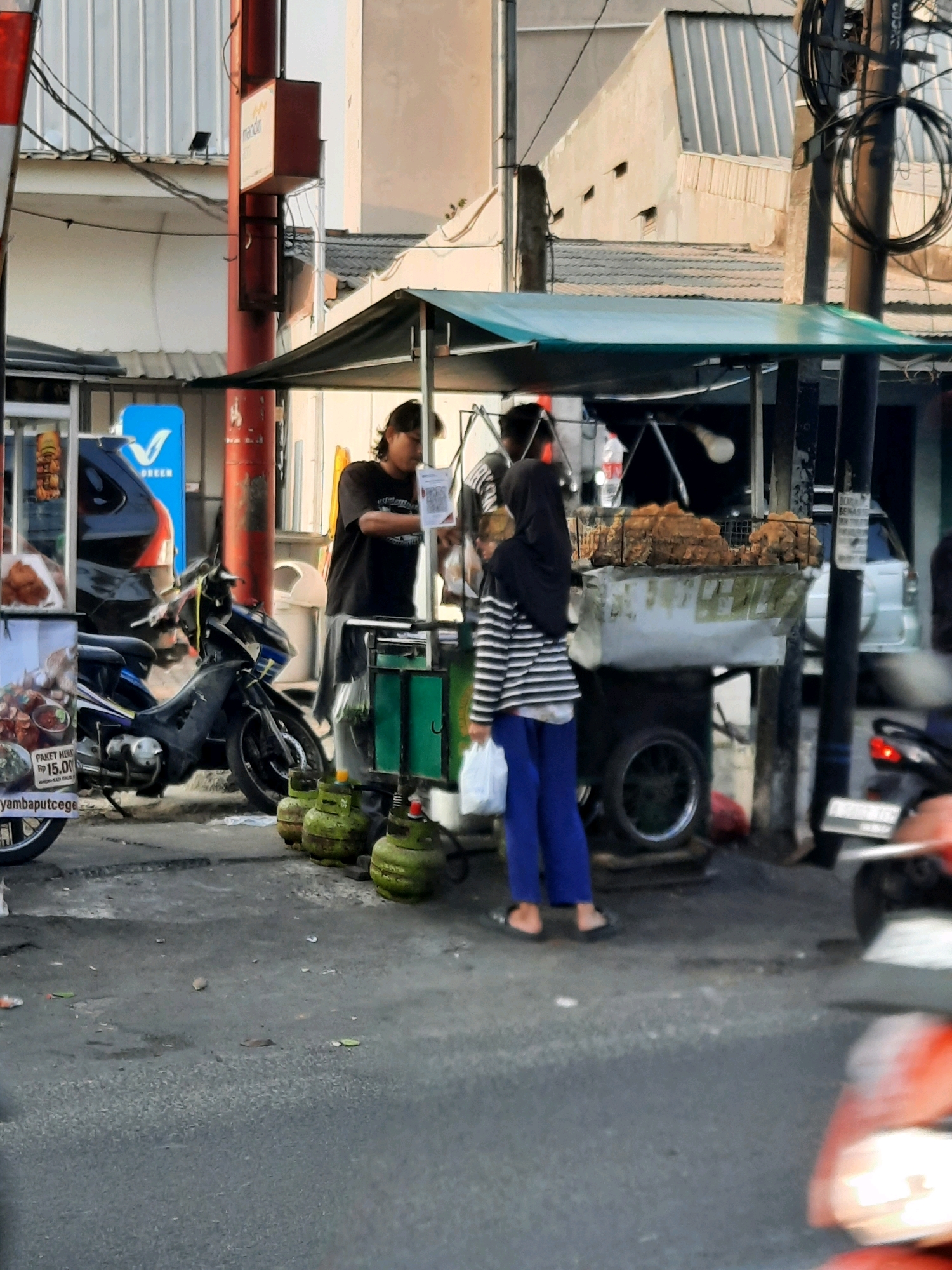 This image depicts a street scene in East Jakarta, Indonesia, likely during daylight. In the foreground, a black and blue motorcycle is parked on a paved street, next to a food cart with a green awning. Three green propane tanks are lined up in front of the cart. To the left of the motorcycle, another food cart displays images of food and prices, with text indicating "PAKET HEMAT RP 15.00".
Behind the motorcycle, a man in a black t-shirt is standing near the food cart, looking down at something. Further to the right, a woman wearing a black hijab and a striped long-sleeved shirt and blue pants is holding a white plastic bag and standing near the food cart. The food cart itself has a white covering with what appears to be fried food on top.
In the background, buildings with corrugated metal roofs and light-colored walls are visible. A red pole with a red box sign featuring the word "mandiri" is also present. Electrical wires crisscross overhead. To the far right, a white car is parked, and parts of a red motorcycle are visible in the foreground, indicating ongoing traffic or parked vehicles. The overall scene suggests a bustling commercial area, possibly a marketplace or street food vendor zone.
