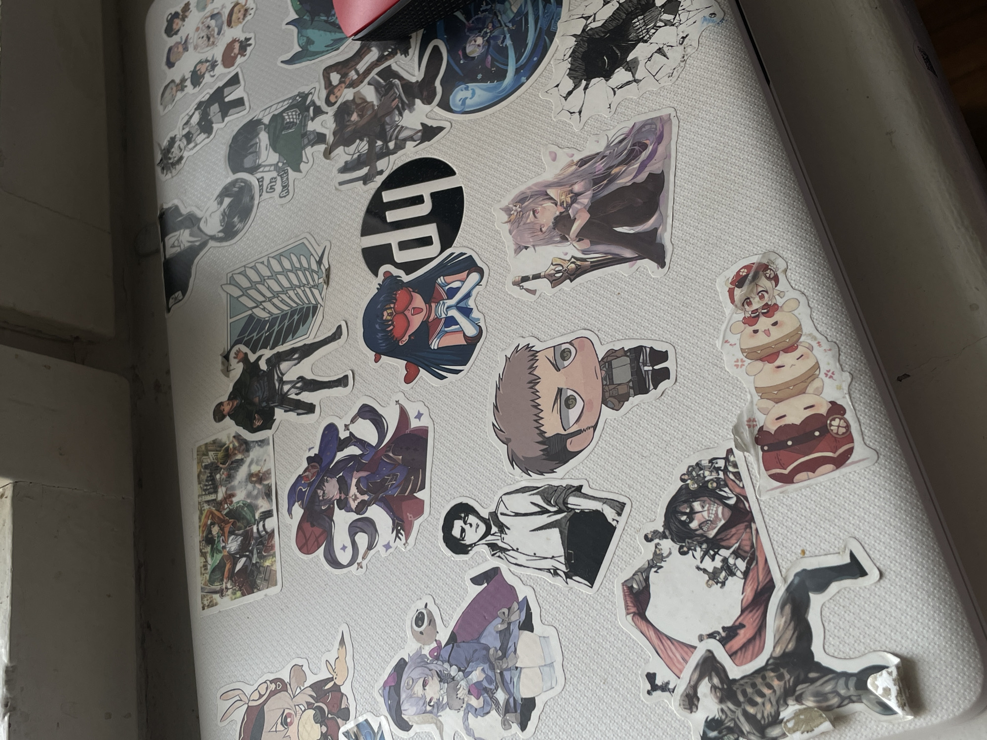 The image displays a close-up, high-angle shot of a laptop lid extensively covered with anime and manga-themed stickers. The laptop itself is light-colored, possibly white or light grey, and features a subtle textured surface. A prominent black circular sticker with the "HP" logo is visible near the center of the lid.
The stickers showcase a diverse range of characters. Several characters from *Attack on Titan* are identifiable, including Mikasa Ackerman, Levi Ackerman, Jean Kirstein, and Eren Yeager (both in human and Attack Titan forms). The "Wings of Freedom" emblem from the series is also present. From *Genshin Impact*, characters like Keqing, Mona, and a chibi-style Klee (perched on stacked items) can be seen. Another notable sticker features Sailor Mars from *Sailor Moon* wearing sunglasses. The stickers are arranged in a dense, overlapping fashion, creating a personalized and colorful surface.
Part of a red computer mouse is visible at the top edge of the frame. The lighting suggests it is daytime, with soft shadows indicating an indoor setting, likely a room, with the laptop resting on a flat surface such as a desk or table. The background is blurred, showing plain wall or furniture surfaces. The overall scene reflects a personal expression of fandom and interest in anime and manga.
The only visible text is the "HP" logo.