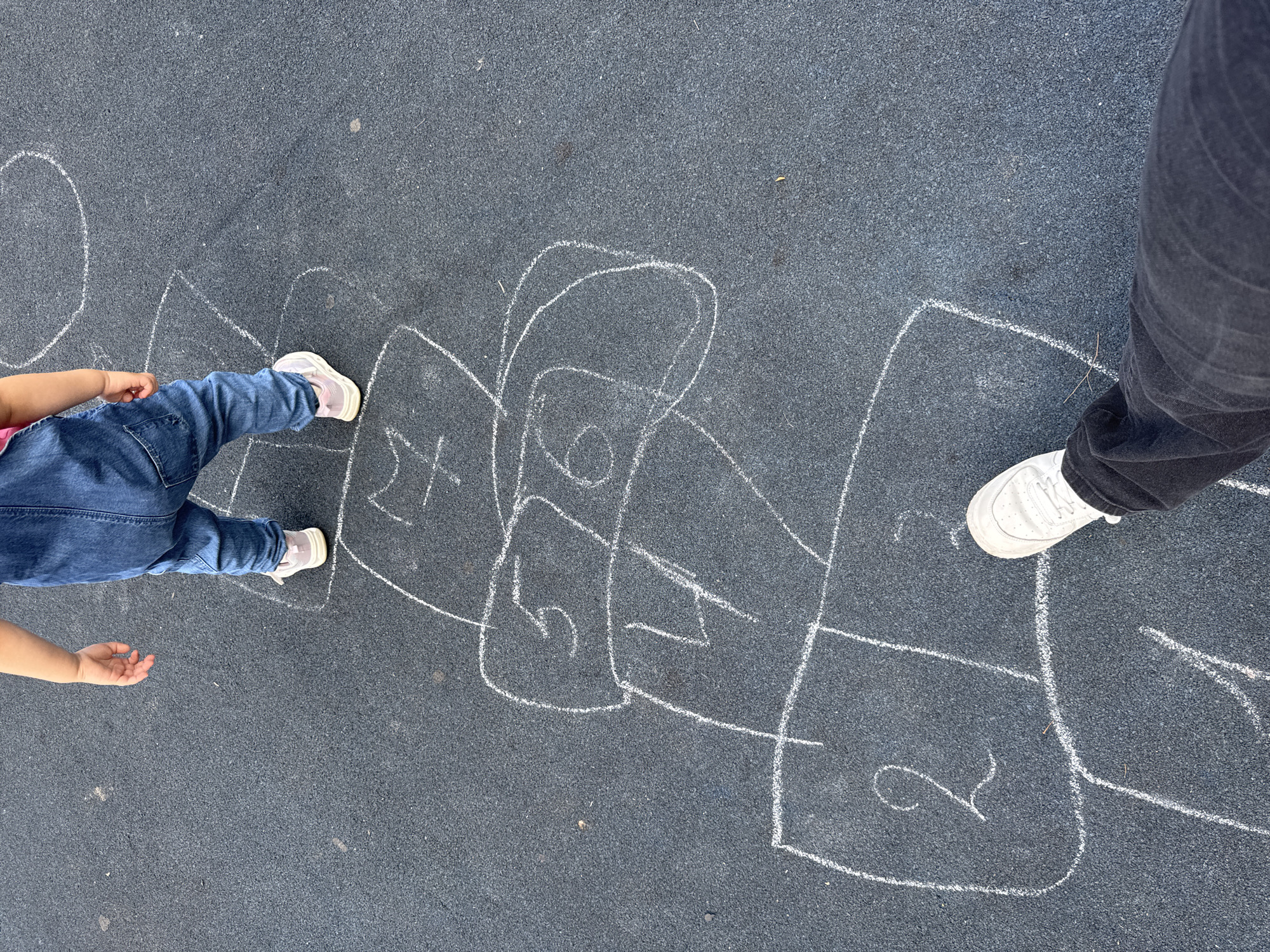 The image shows two people playing hopscotch drawn with chalk on an asphalt surface. The person on the left, a child, is wearing blue jeans and a pink top and is mid-stride with their left leg extended, wearing white sneakers with pink accents. Their right arm is extended and their hand is open. The person on the right is an adult wearing dark pants and white sneakers. The hopscotch court is drawn with white chalk and includes numbered squares. The numbers visible are "6", "4", and "2". The scene appears to be outdoors in daylight, as indicated by the shadows and the brightness of the image. The setting seems to be a playground or a paved outdoor area. The activity taking place is a game of hopscotch, suggesting a playful and recreational atmosphere. The image is taken from a top-down perspective, focusing on the feet and lower legs of the players and the hopscotch court.