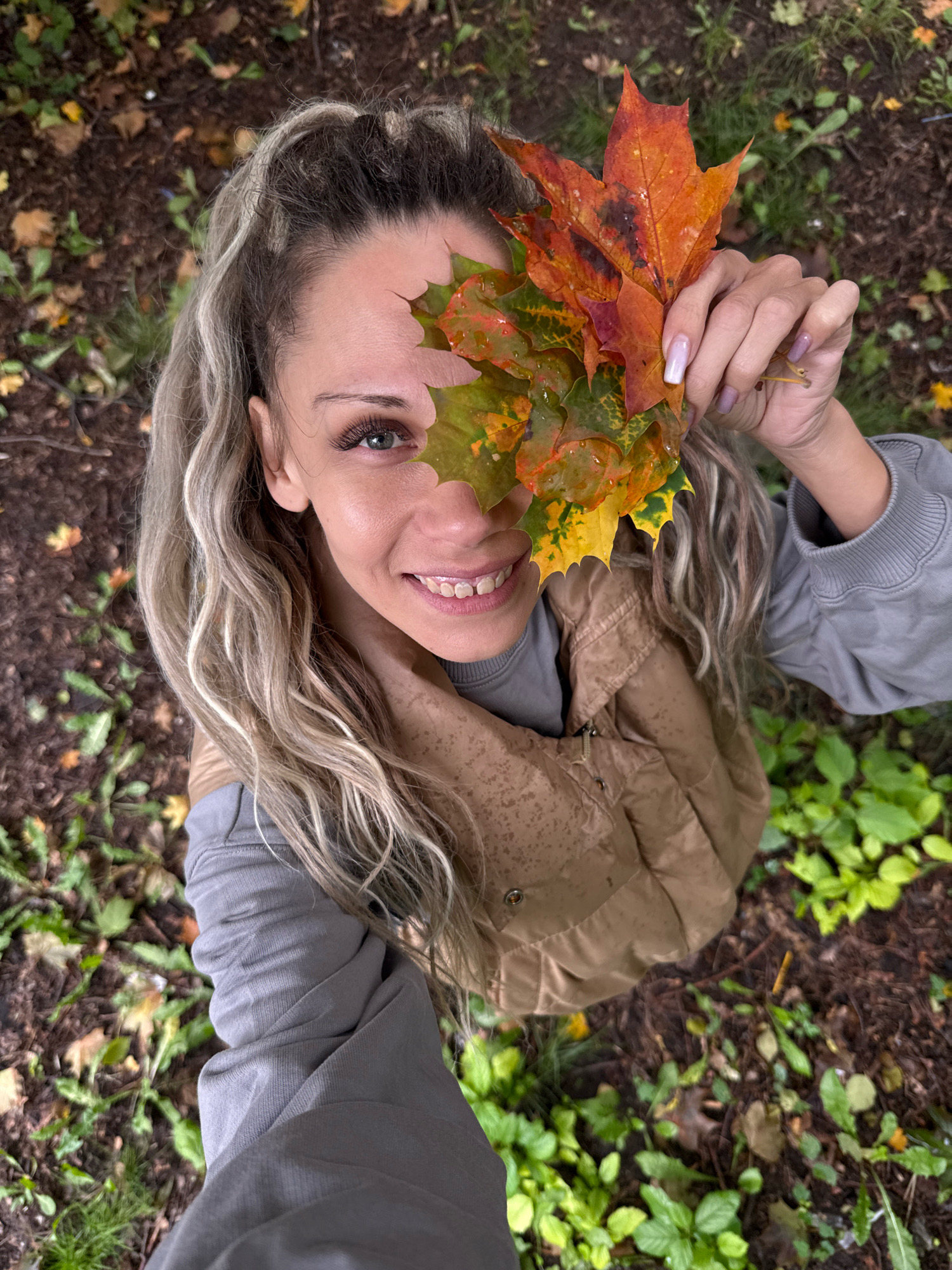 A high-angle, medium shot captures a woman holding a bunch of colorful autumn leaves in front of her face, obscuring her eyes and nose. She is wearing a light gray crewneck sweatshirt layered under a brown puffer vest. Her blonde hair is pulled back in a ponytail, with several strands framing her face. Her right hand, with a light pink manicure, holds the leaves. She is smiling, with her mouth open to reveal her teeth. The background consists of a forest floor covered in brown leaves and green vegetation. The scene is set in Rusanovo, Russia, during autumn.