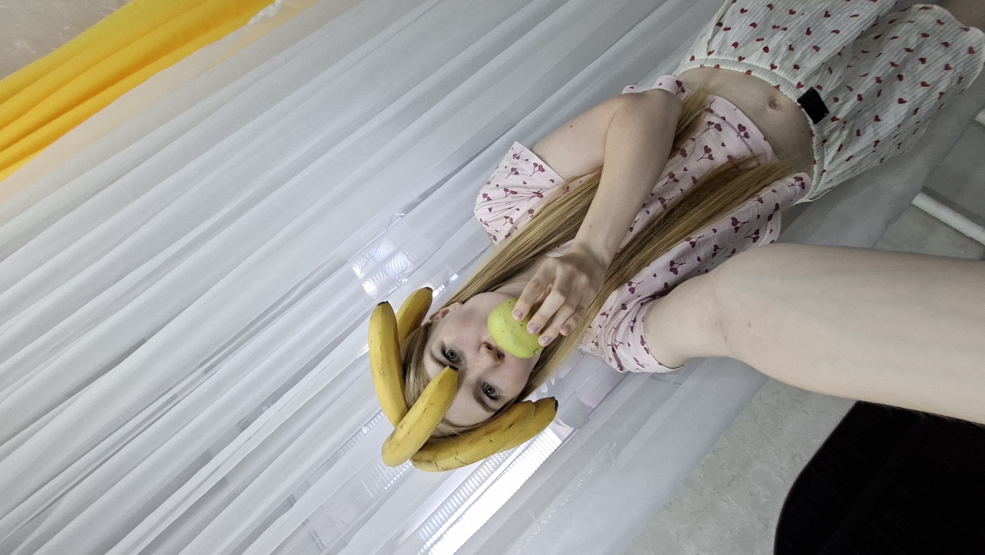 The image features a young woman with long, straight blonde hair, light skin, and blue eyes, positioned in a playful, reclined pose. She is looking directly at the camera. A cluster of ripe yellow bananas is artfully arranged on her head, resembling a decorative crown. She holds a light green apple near her mouth with her right hand, suggesting she is about to take a bite or is posing with it.
The woman is dressed in light-colored clothing; her top features a pink and white pattern, possibly of small hearts or cherries, and she appears to be wearing matching shorts or bottoms, exposing her midriff.
The background is predominantly composed of white, translucent fabric, likely sheer curtains or drapes, which provide a soft, diffused light. In the upper left corner, a vibrant yellow fabric is visible, possibly another layer of curtain or a decorative element, adding a pop of color to the otherwise muted background. The image appears to be taken indoors during the daytime, given the bright, ambient lighting. No text is visible in the image. The overall mood is whimsical and relaxed. The location context is Chaikovsky, Russia.