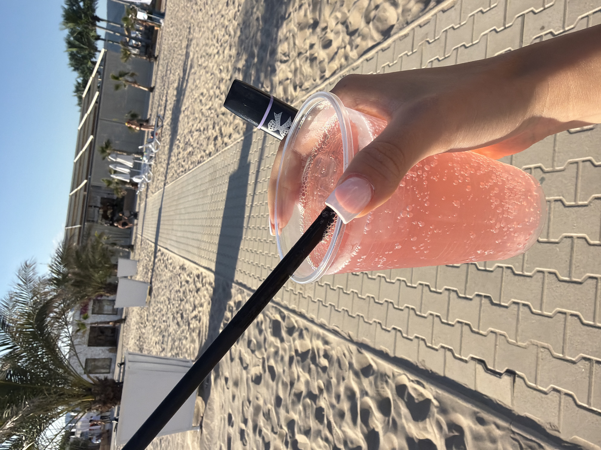 A close-up shot captures a person holding a clear plastic cup filled with a pink, bubbly beverage. A black straw extends from the drink. Resting atop the cup is a small, black object with a light purple band and a white bow design, possibly a vape pen or a lipstick. The person's hand, with neatly manicured nails, is visible as they grip the cup.
The background suggests a sunny outdoor setting, likely a beach resort. A paved pathway made of light-colored interlocked bricks runs diagonally across the image. To the left of the pathway, fine sand is visible, marked with footprints and indentations. Palm trees and white planters are scattered across the sandy area. Further in the background, a building with a white facade, possibly a beach bar or restaurant, is visible, with people and lounge chairs nearby. The sky is a clear, bright blue, indicating fair weather. The scene appears to be located in a resort area, possibly on the coast of Nesebar, Bulgaria, given the typical architecture and palm trees found in such locations.