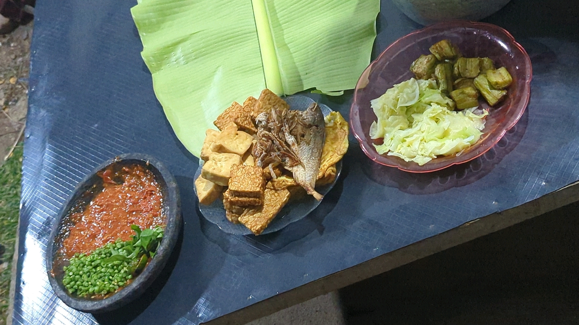 In Semut, Indonesia, a close-up overhead shot captures a rustic meal laid out on a dark, textured surface, possibly a wooden table. The scene is dominated by three plates of food. To the left, a dark, oval-shaped stone mortar holds a vibrant red sambal, a spicy chili paste, adorned with fresh green peas and herbs. In the center, a clear glass plate overflows with a variety of fried items, including cubes of golden-brown tofu, rectangular pieces of tempeh with visible fermentation pores, and a whole fried fish with its head intact. Beside the fish, a few pieces of what appear to be fried omelets are visible. To the right, a pink plastic bowl contains a side dish of shredded cabbage and what seems to be pieces of cooked eggplant. A large, vibrant green banana leaf serves as a natural placemat or backdrop for the central plate. The lighting suggests it might be evening, casting subtle shadows. The ground to the left of the table appears to be gravel or dirt.