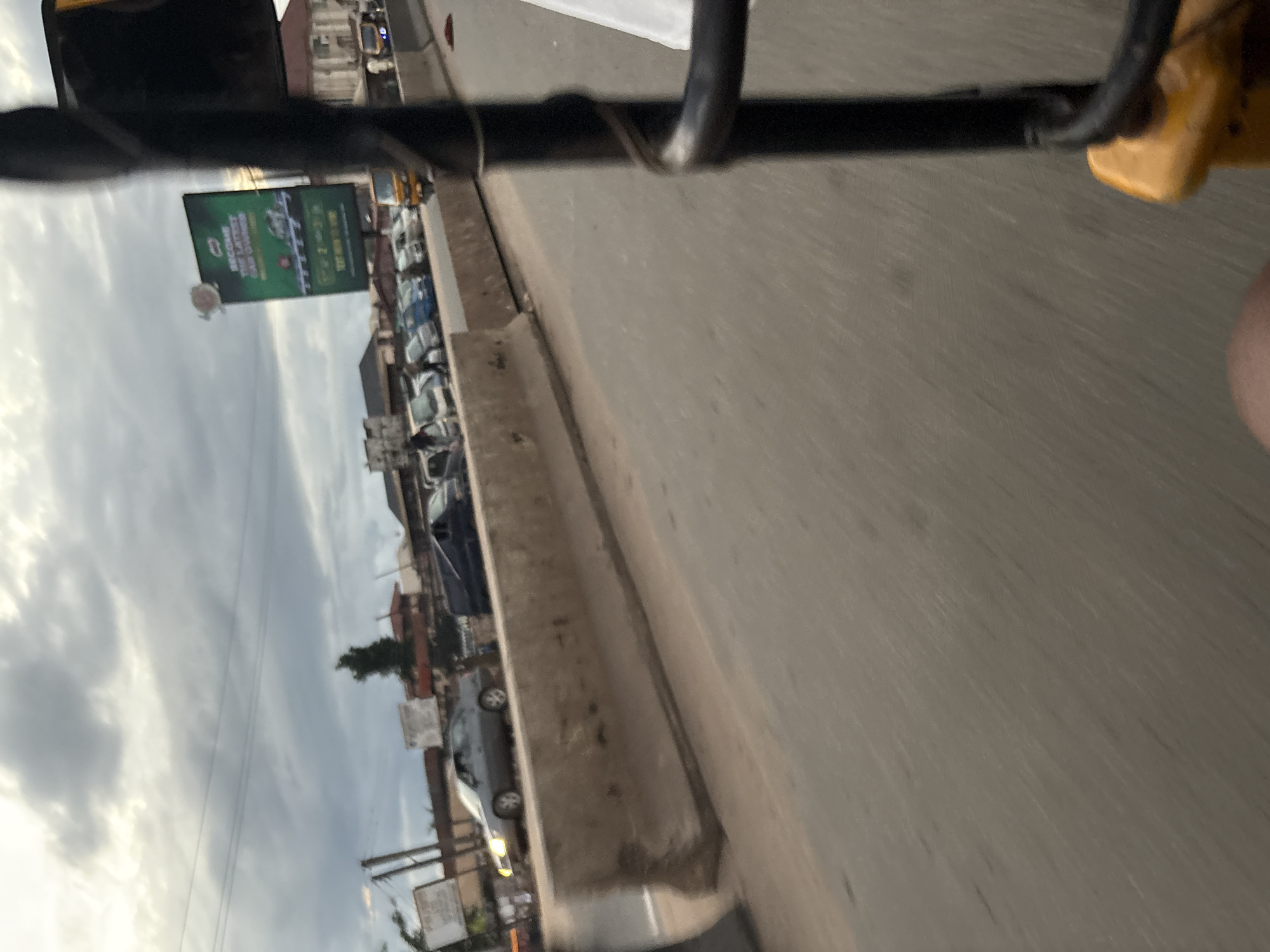 This image, captured from a moving vehicle, depicts a street scene in Onitsha, Nigeria. The foreground is dominated by the dark frame of the vehicle, with a yellow seat visible at the bottom right. A concrete barrier runs along the side of the road, separating it from a line of parked or slow-moving vehicles including a silver car with its headlights on, several blue cars, and a yellow tricycle. In the background, buildings line the street under a cloudy sky. A large green billboard advertising "BECOME THE LATEST CAR OWNER" is prominently displayed on the right side of the image. The overall impression is one of a busy urban environment.