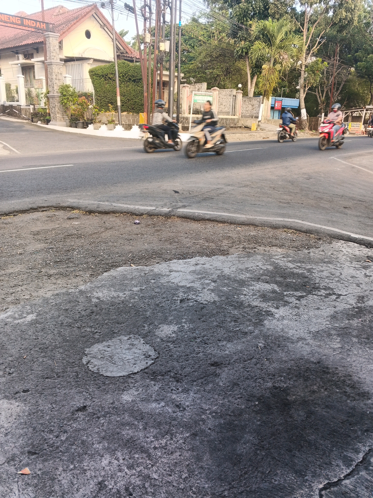 The image shows a street intersection in Semarang, Indonesia, with several people riding motorcycles. In the foreground, there is a damaged section of asphalt road, with a pothole filled with gravel and dirt. In the background, a building with a red-tiled roof and a sign reading "NENG INDAH" is visible. There are also several utility poles and lush green trees lining the street. The sky is overcast. The people on the motorcycles are blurred due to motion.