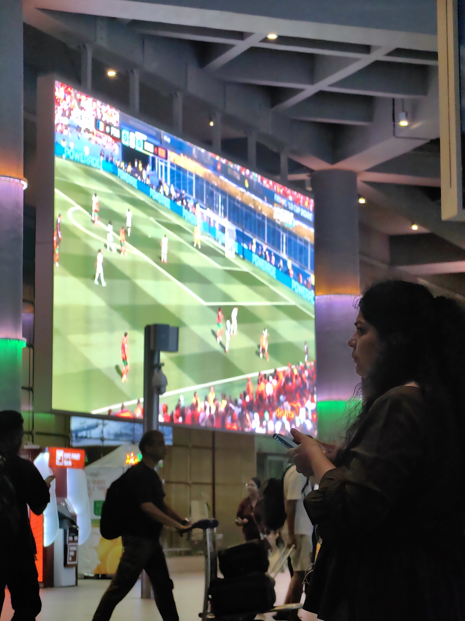 The image captures a large, bright screen displaying a soccer match, likely in a public space like an airport or transit hub. The screen shows players in red and white uniforms on a green field with spectators in the background. In the foreground, a woman is seen from the side, looking towards the screen while holding a smartphone. She has dark, curly hair pulled back. To her left, other individuals are visible, including a man with a backpack and a person in a black shirt pulling a trolley. The environment appears to be indoors, with overhead structures and lighting. The presence of a large screen showing a sporting event suggests a place where people gather to watch or wait. The image was taken in Mumbai, India.