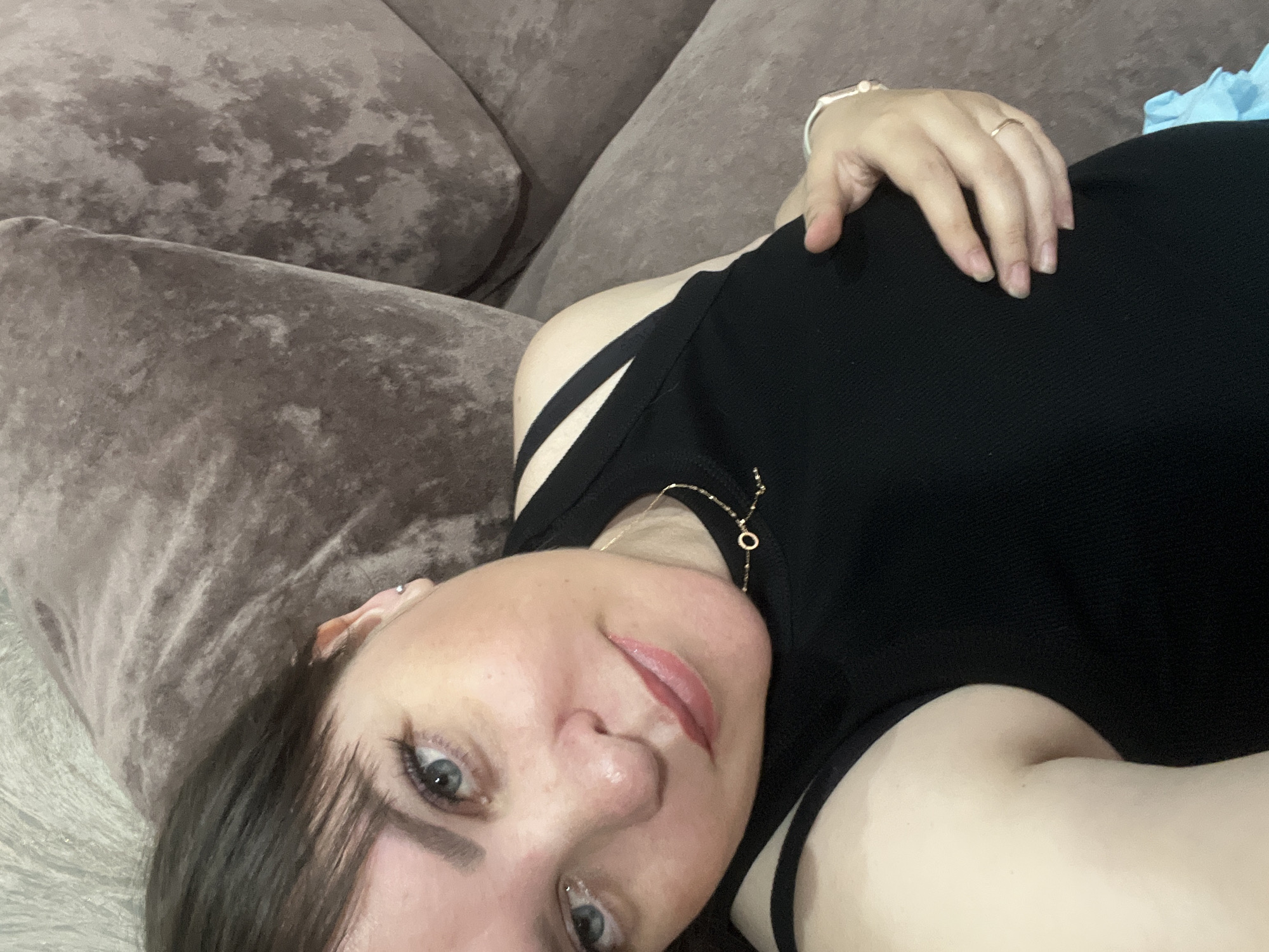 A person with fair skin and short, dark hair lies on a brown, textured couch. The person wears a black top with strap detailing and a delicate gold necklace. Their left hand, adorned with a ring and a watch, rests on their torso. The photograph's perspective is angled, focusing on the person's face and upper body. No information exists in the image indicating a specific location in Noviy, Russia.