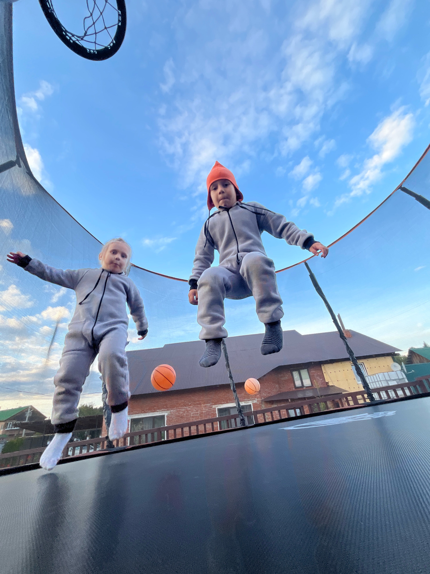 In the city of Chaikovsky, Russia, two children are captured mid-air on a trampoline against a backdrop of a bright blue sky with scattered clouds. The child on the left, a girl wearing a gray hooded jumpsuit and white socks, extends her arms outward. The child on the right, a boy in a gray hooded jumpsuit and an orange beanie, is also in mid-jump, his left leg bent and his right leg extended. Two orange basketballs are visible, one near the girl and another further to the right. Behind the trampoline, houses with brick and light-colored exteriors, a wooden fence, and trees are visible. The trampoline has a black mat and netting. The low-angle perspective emphasizes the upward motion of the children.
