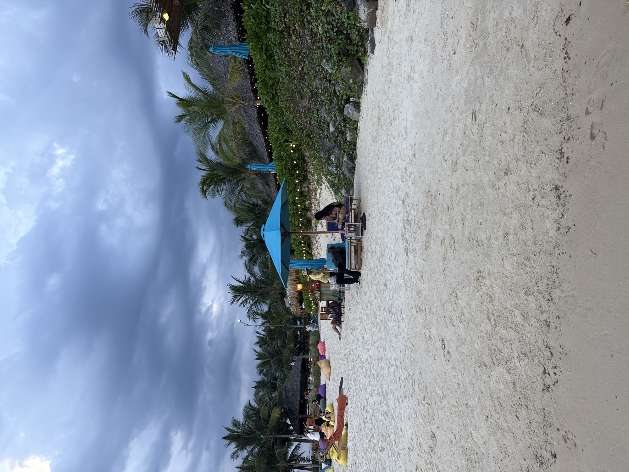 The image shows a beach scene in Bac Nha Trang Ward, Vietnam. A wide sandy beach occupies much of the foreground and right side. Along the left side of the frame, palm trees line the beach, with some buildings and thatched structures interspersed. String lights hang from the structures, adding a warm glow to the scene. Several blue beach umbrellas are visible, shading seating areas on the beach. People are present, some sitting on chairs and cushions. The sky is overcast, filled with dark, heavy clouds.