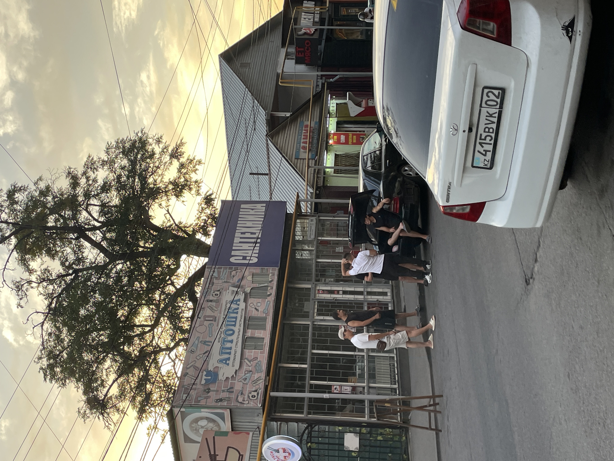 The image depicts a street scene in Almaty, Kazakhstan, during the late afternoon or early evening, as indicated by the warm, diffused sunlight and soft shadows. The foreground is dominated by the rear of a white Chevrolet Gentra sedan, with a license plate reading "KZ 415BVK 02". A black car with its trunk open is parked on the street behind the Gentra.
Several men are present in the scene, appearing to be engaged in conversation or socializing. One man, wearing a white t-shirt and beige shorts, stands on the sidewalk. Another man in a black tank top and shorts leans against the black car. Two other men, one in a white t-shirt and the other in black clothing, are also visible near the cars.
The background reveals a row of commercial establishments. One building features signage for "АПТОШКА" (Aptoshka), which appears to be a hardware or construction materials store, with an illustration of tools on its facade. Adjacent to it, another sign reads "САНТЕХНИКА" (San-tekhnika), indicating plumbing supplies. Further down the street, more shops are visible, including one with a sign that seems to say "МЯСО" (Myaso), meaning meat. The buildings are of varying architectural styles, some appearing to be single-story structures with corrugated metal roofs.
Overhead, a network of electrical wires crisscrosses the sky, typical of urban infrastructure. A large tree with dense foliage stands prominently in the upper left portion of the image, its branches silhouetted against the hazy sky. The overall impression is a snapshot of everyday life on a commercial street in Almaty, capturing a moment of informal gathering and commerce.